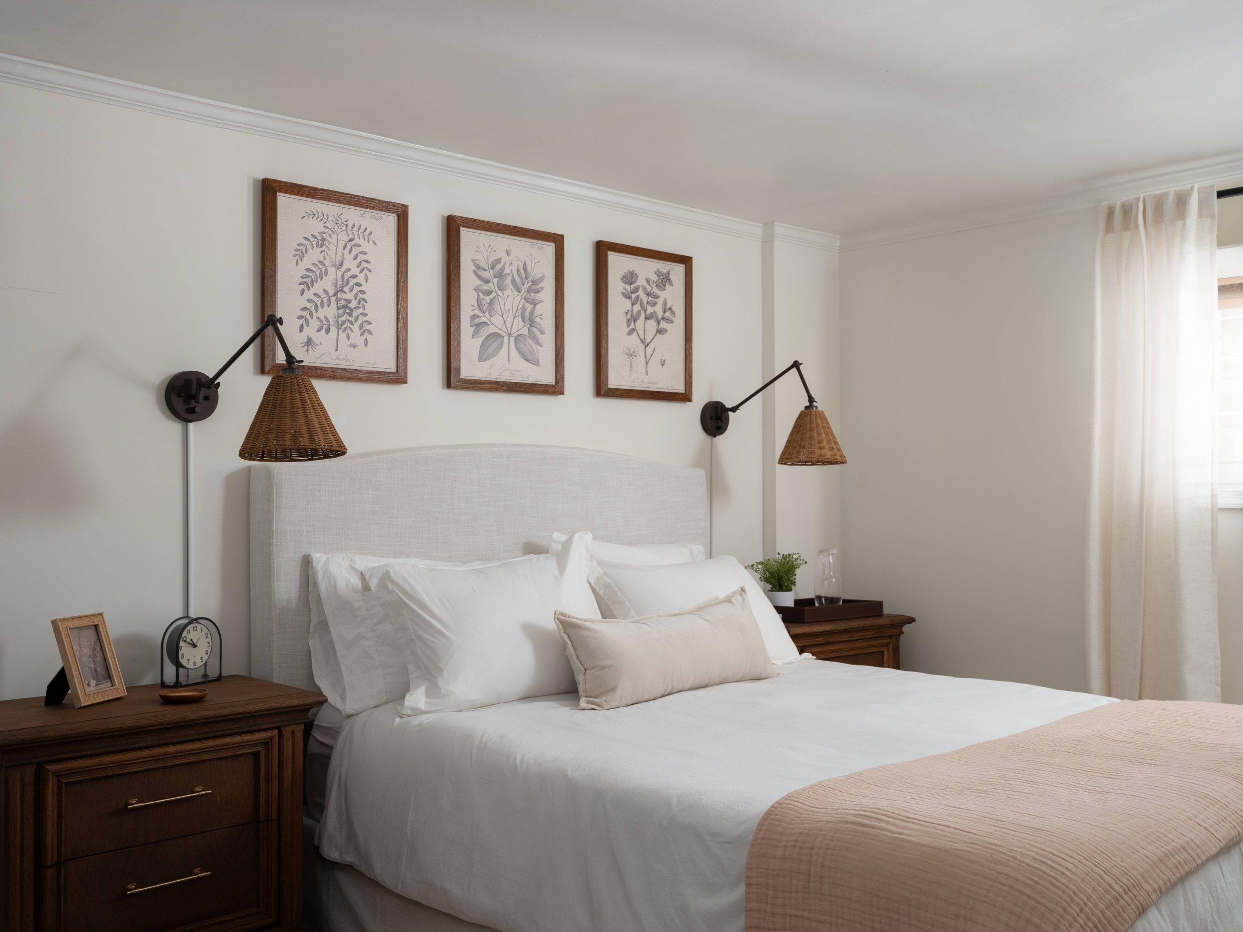 A cozy bedroom with a large bed dressed in white linens, resting against a padded white headboard. The wall behind features three framed botanical prints, and two matching black wall-mounted lamps with woven rattan shades flank the bed. A wooden nigh