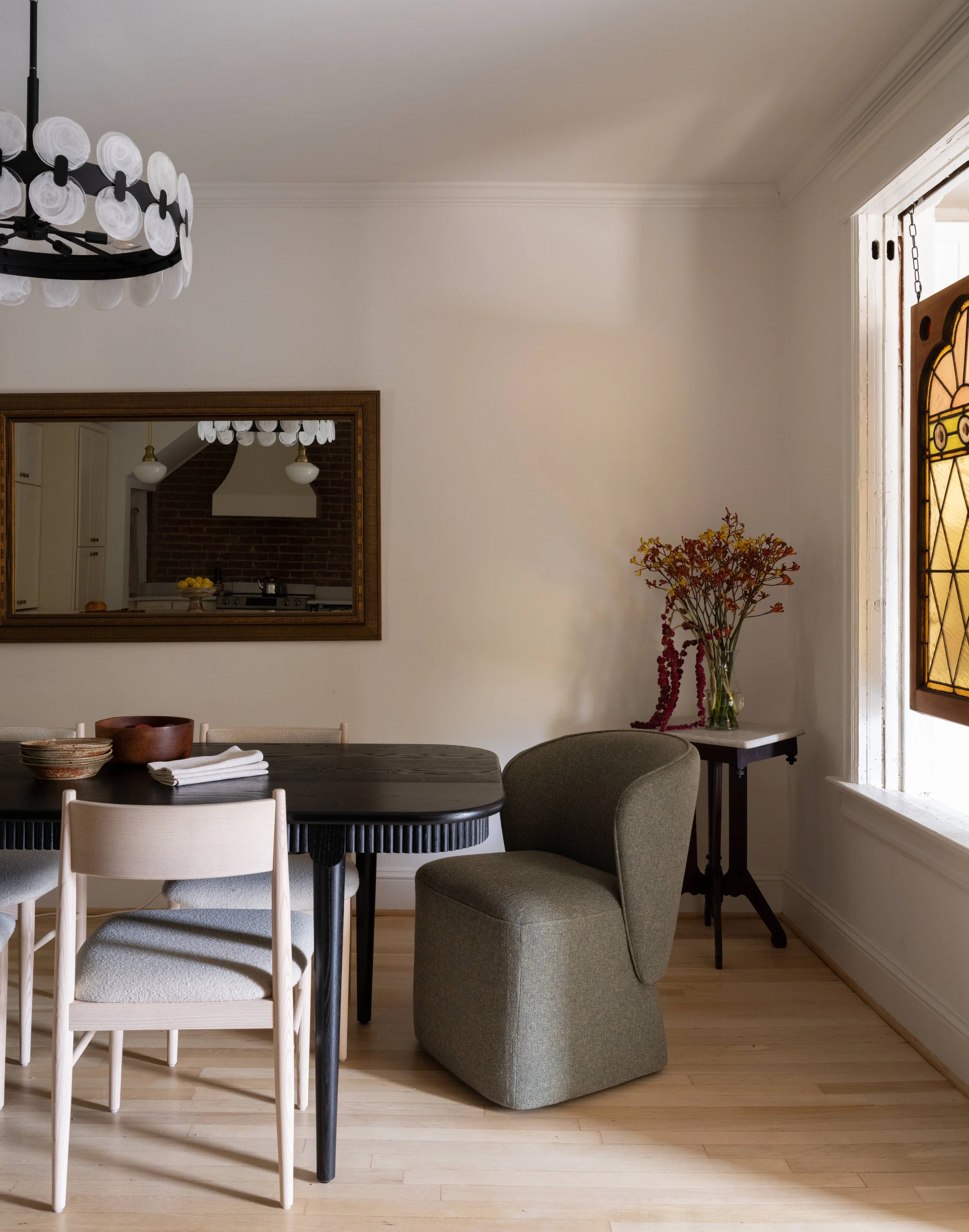 Dining room with a black table, white chairs, a gray upholstered armchair, a side table with a vase of flowers, a large mirror, a wall chandelier, and a window with stained glass.