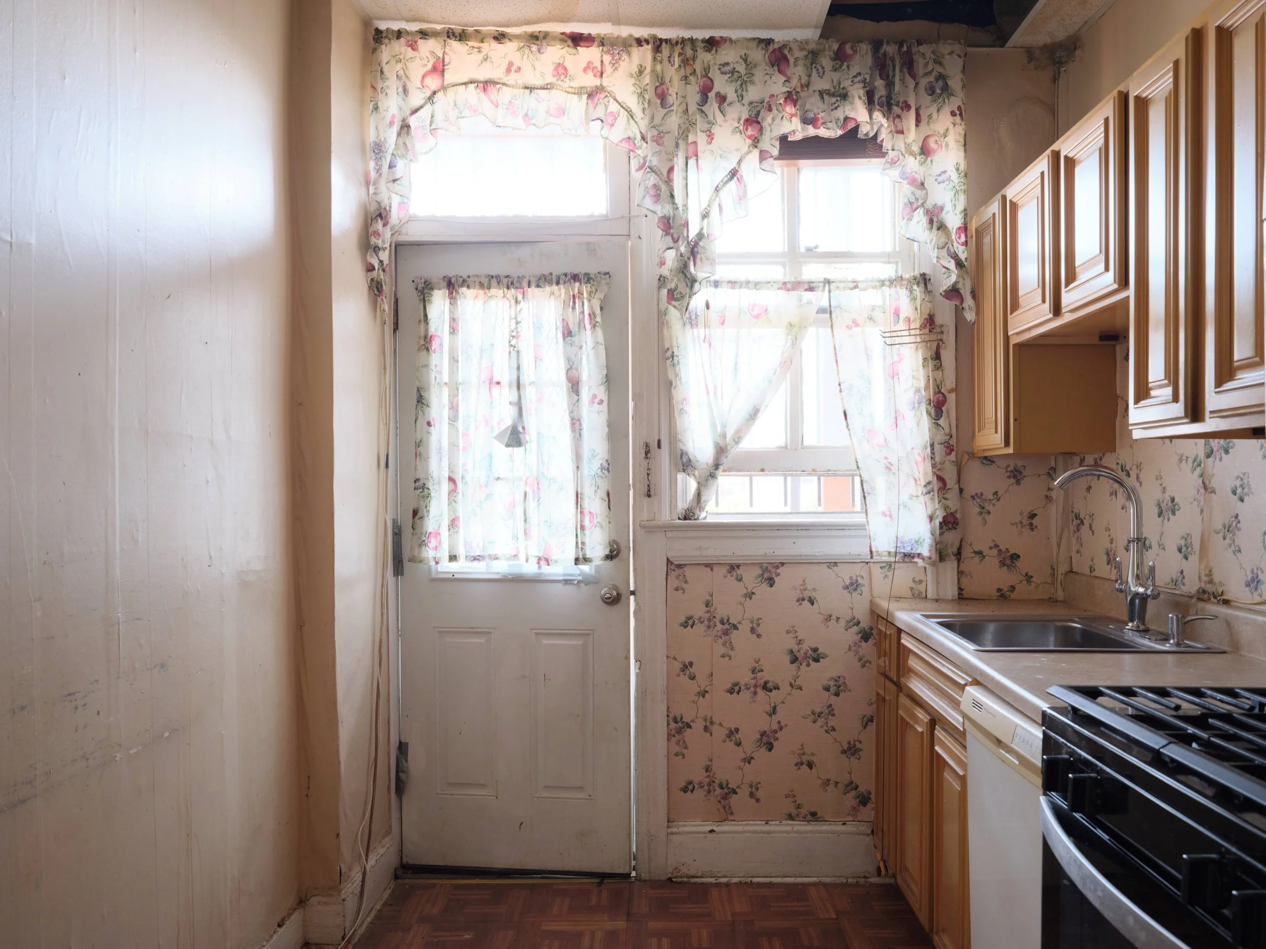 A small kitchen with floral curtains on a window and a door, wooden cabinets on the right, a sink, and a stove. The walls and ceiling are plain and worn.