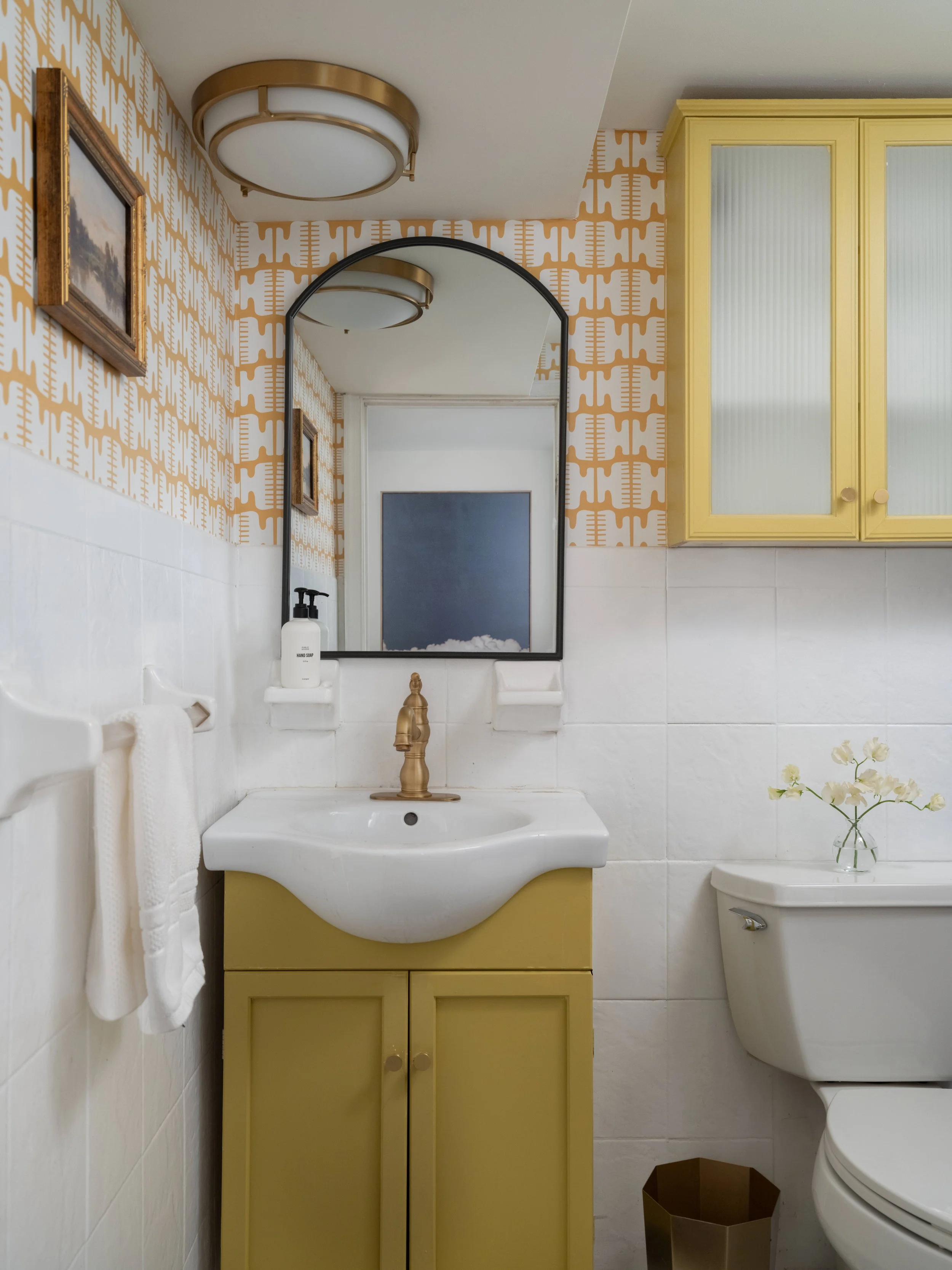 A small bathroom with a yellow cabinet below a white sink, a white toilet, a mirror, a framed painting on patterned wallpaper, a white towel, a soap dispenser, and a small flower vase with flowers.