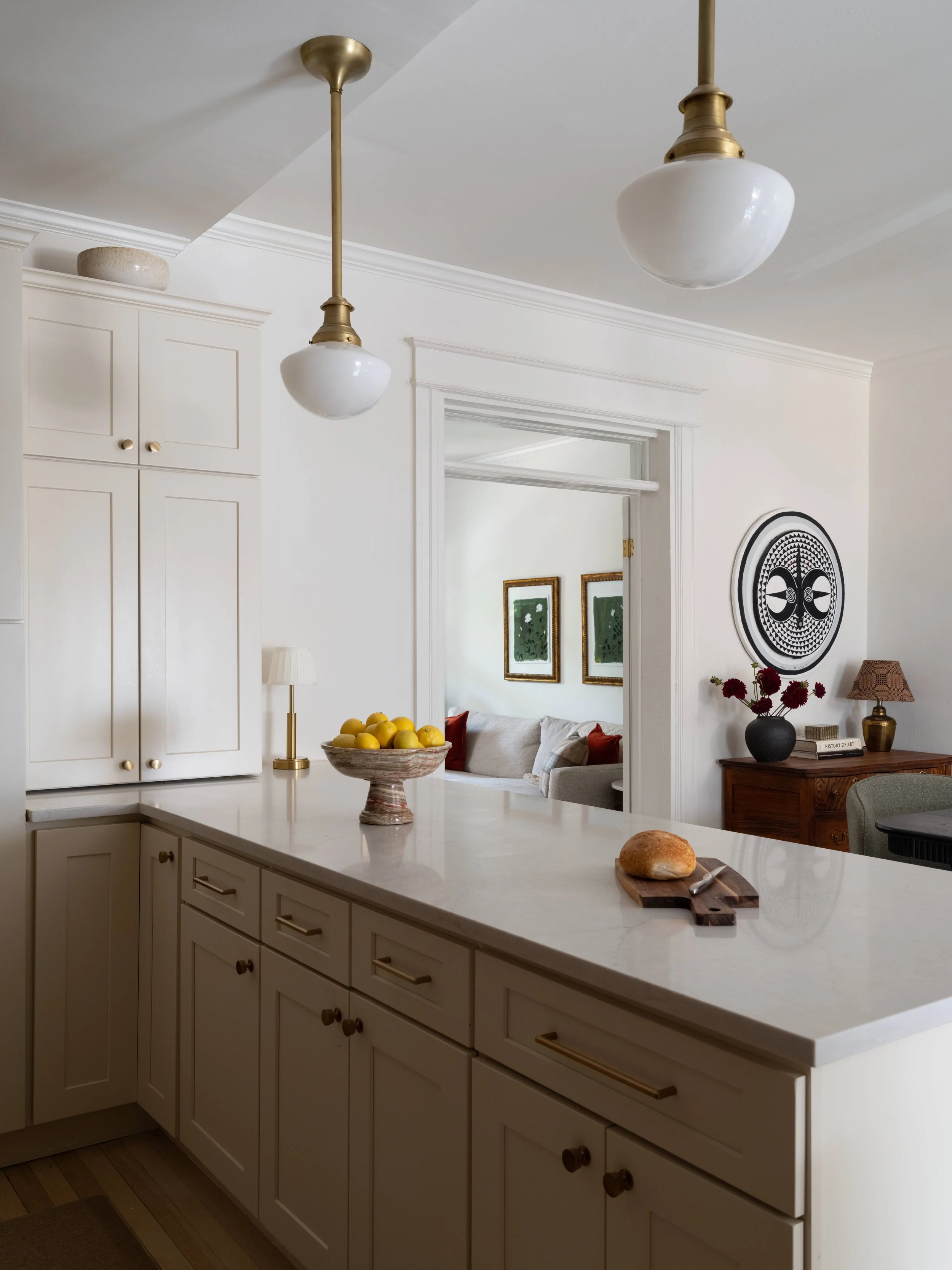 A kitchen with white cabinets and a marble countertop. On the counter, there's a bowl of lemons, a loaf of bread on a cutting board with a knife, and a small lamp. Two white hanging pendant lights with brass fixtures are above. In the background, a l