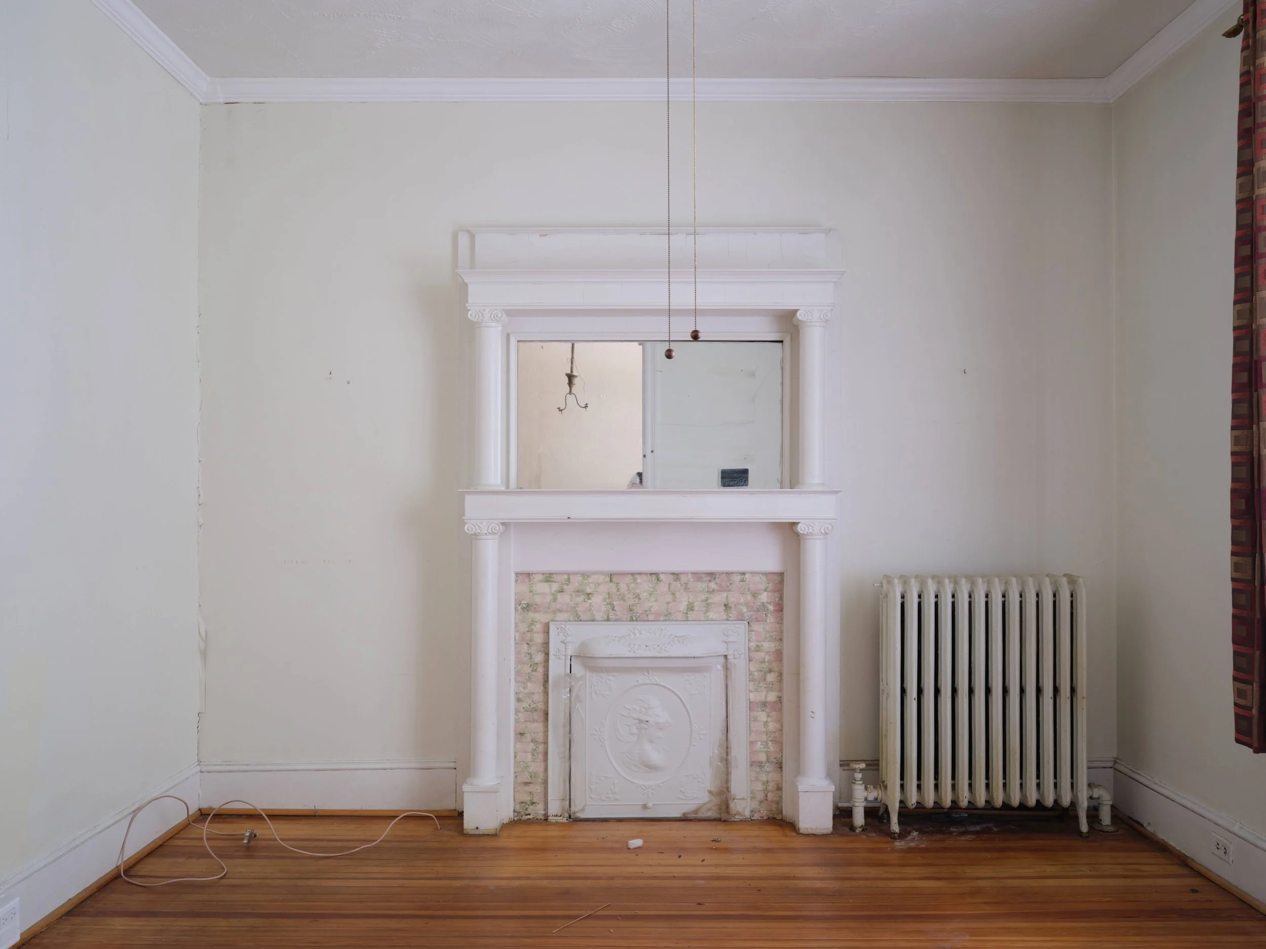 Empty room with a fireplace mantel, mirror above, beige walls, hardwood floor, radiator, and dark red patterned curtains on the right side.