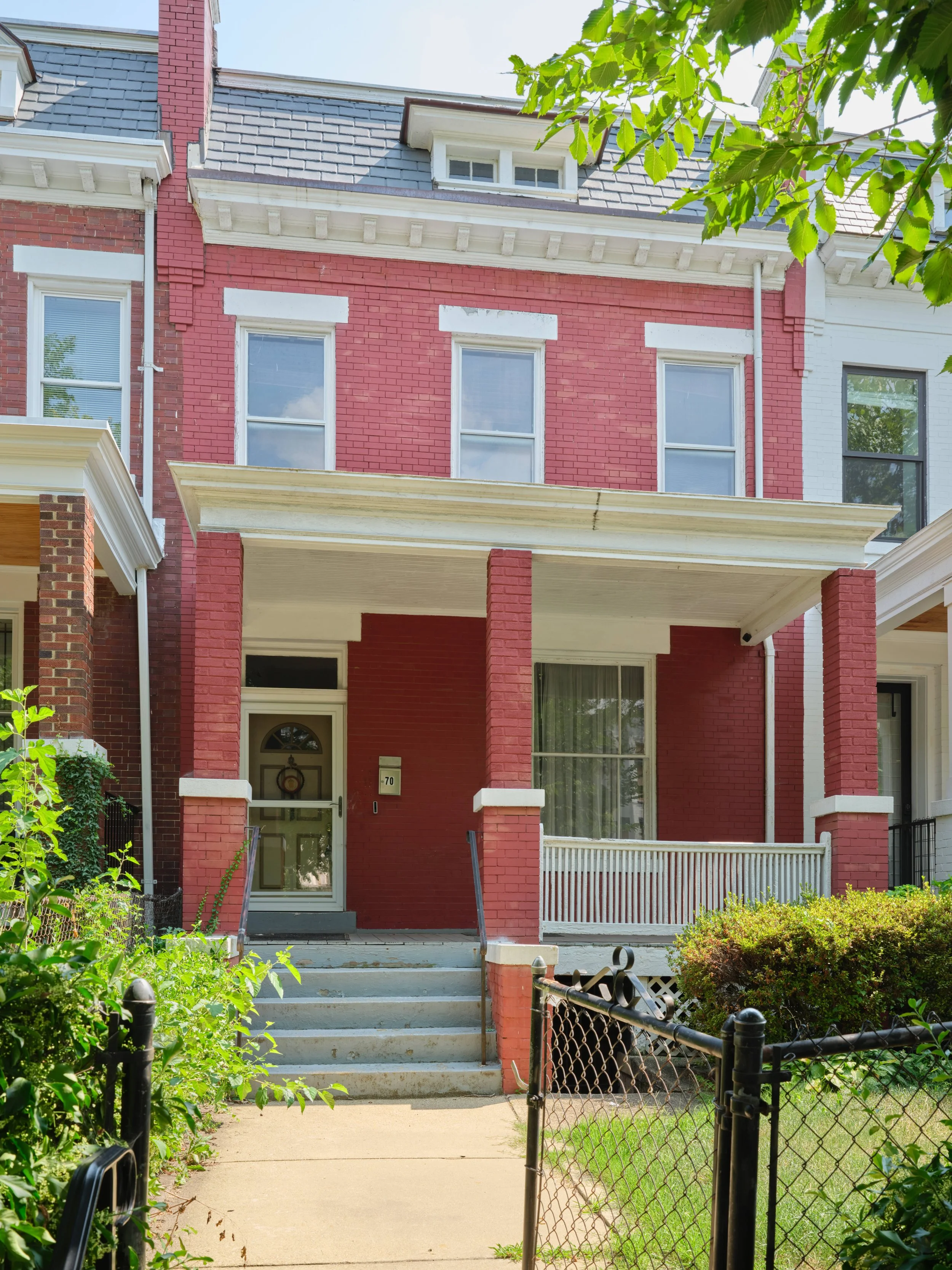 Front view of a red brick house with white trim and a porch, overlooking a sidewalk with green bushes and a black fence.