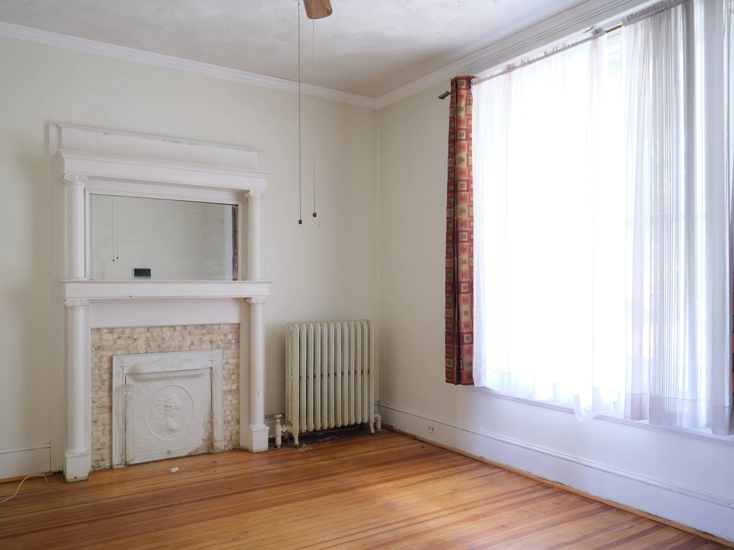 Empty room with wooden floor, white walls, a white decorated fireplace, a radiator, a large window with sheer white curtains and a patterned red and beige curtain, a ceiling fan, and an electrical outlet near the floor.