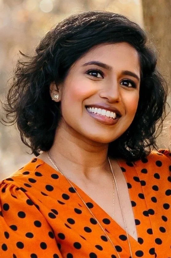 A woman with curly black hair smiling outdoors, wearing a burnt orange dress with black polka dots and a silver necklace.