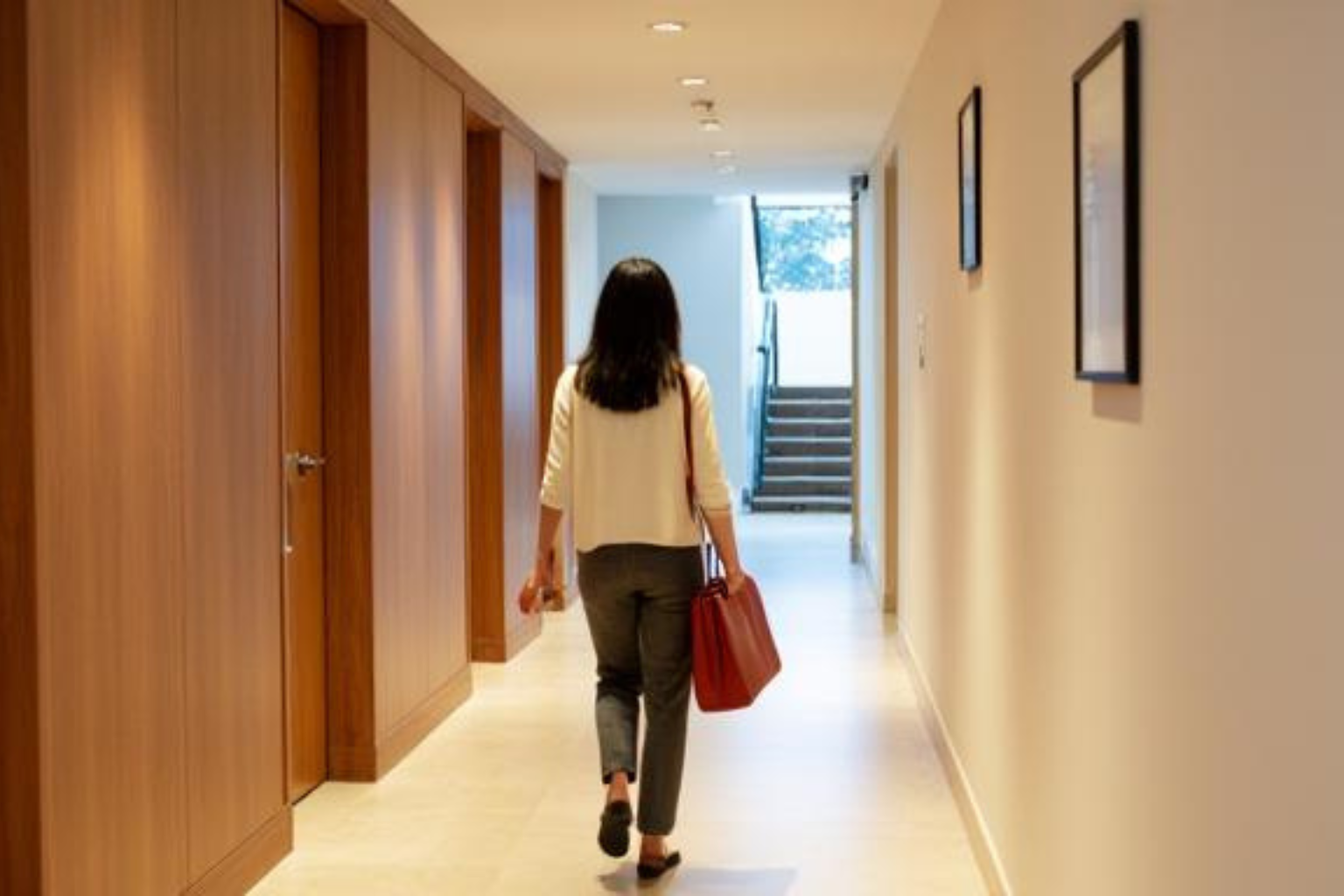 A professional walks alone down a quiet corridor after a meeting, toward a staircase at the far end.