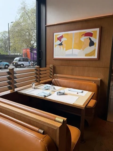 Empty booth seating at a restaurant with a framed abstract art piece on the wall depicting two women wearing hats, and a window showing a street scene outside.