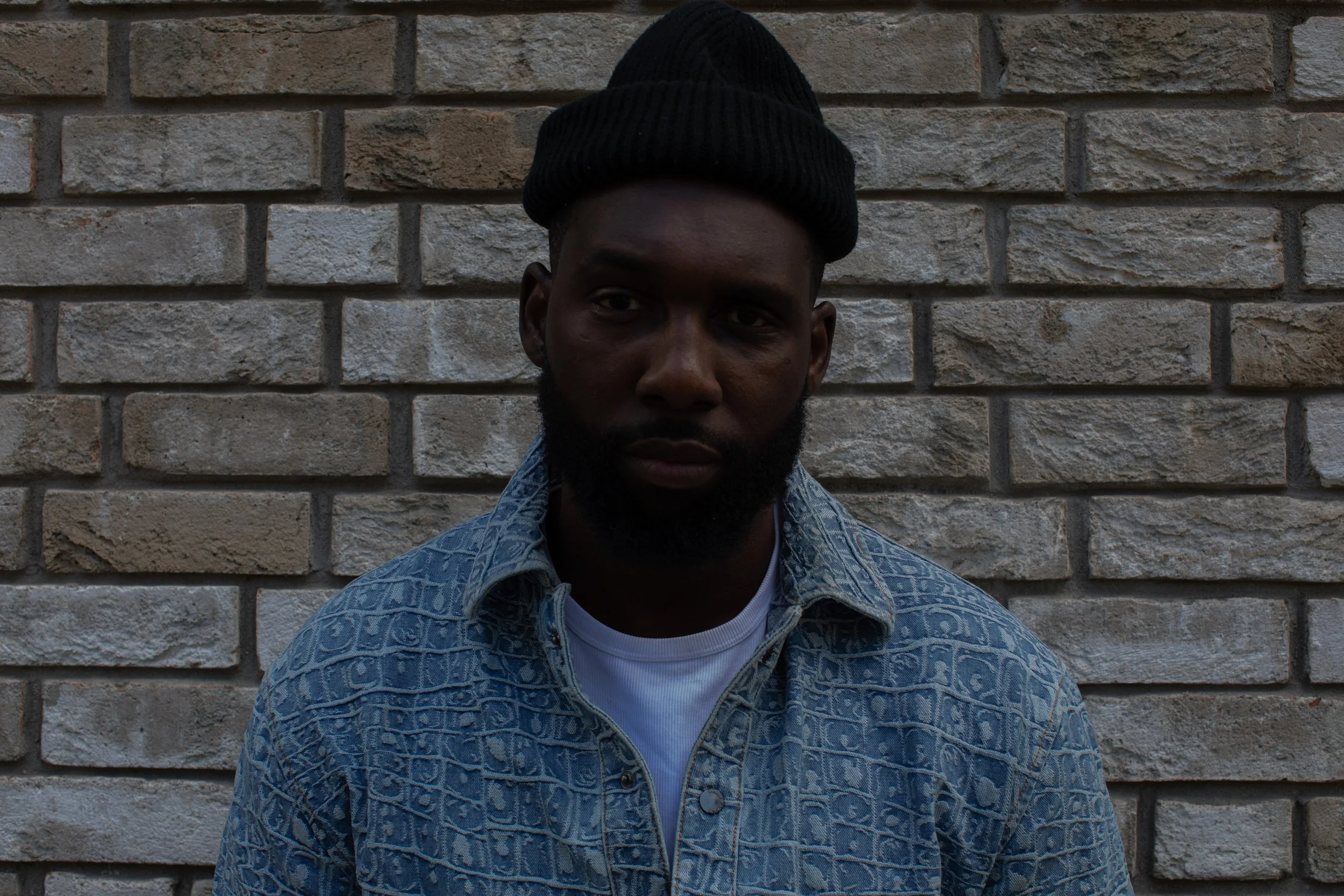 A man with dark skin and a beard wearing a black beanie and a patterned blue denim jacket standing against a brick wall.