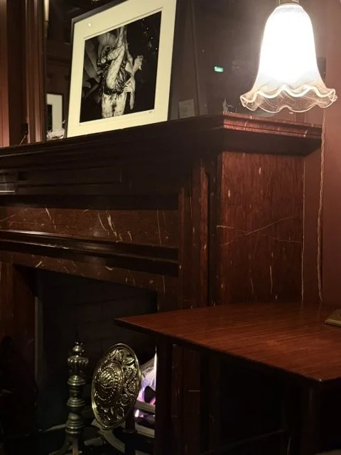 Close-up of a dark wood fireplace mantel with a framed black-and-white photograph on top and a glowing table lamp beside it.