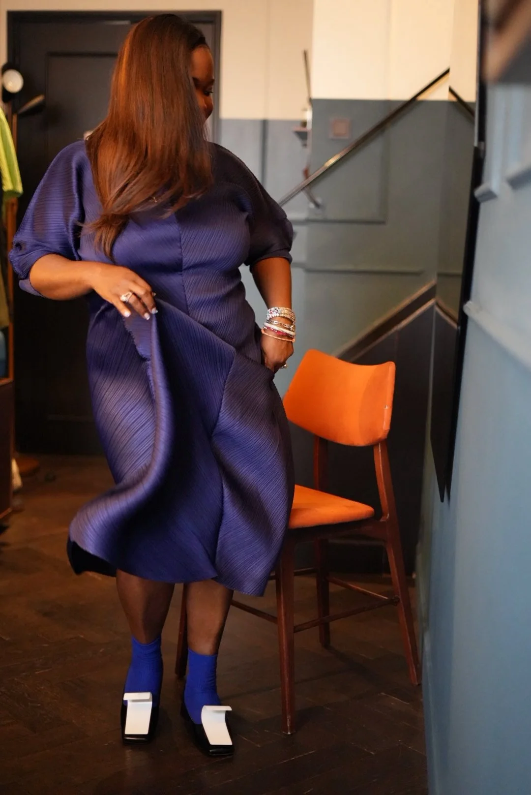 A woman with long brown hair wearing a purple dress, blue socks, and stylish black and white shoes, standing in a room with wooden flooring and an orange chair, adjusting her dress.