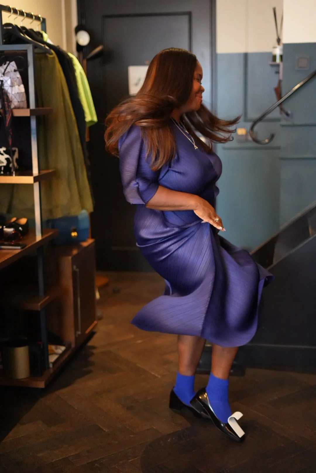 A woman dancing on the stairs in a purple dress, black and patent shoes, and bright blue socks inside a retail store.