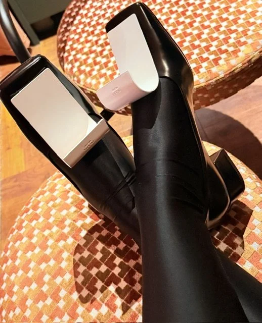 Person lying on a chair wearing black high heels with white soles, with legs crossed and resting on an orange patterned chair seat.