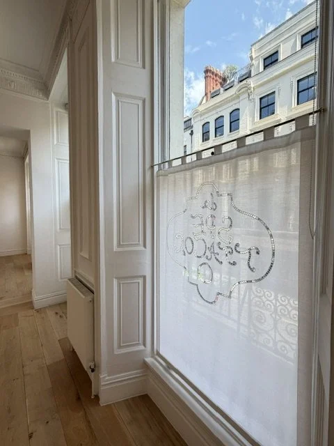View through a window with decorative sheer curtains showing an exterior of a white building with multiple windows and a chimney on top, under a partly cloudy blue sky.