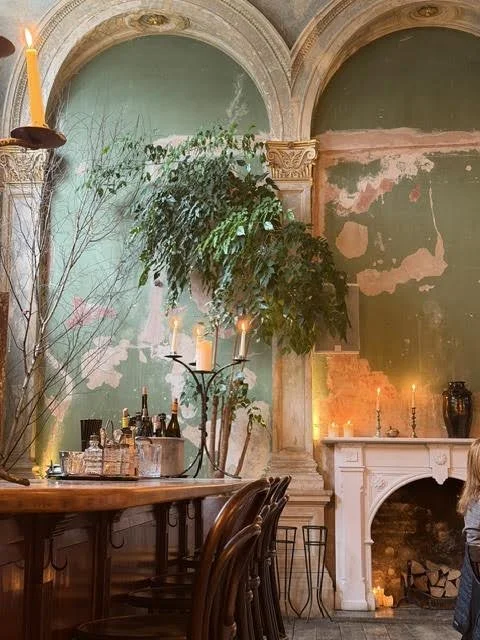 Interior of an old building with distressed green walls, ornate arches, a wooden dining table with chairs, candles, bottles, and a fireplace with candles and decor on the mantel.