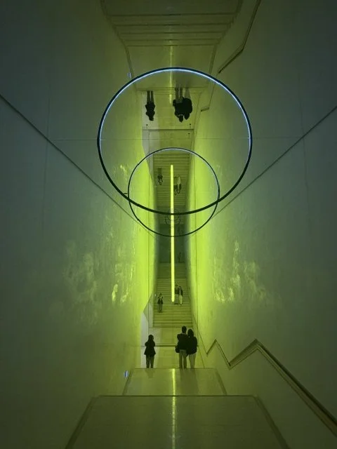 Upside-down view of a stairway with a neon yellow light, two neon circles on the ceiling, and people walking down the stairs.