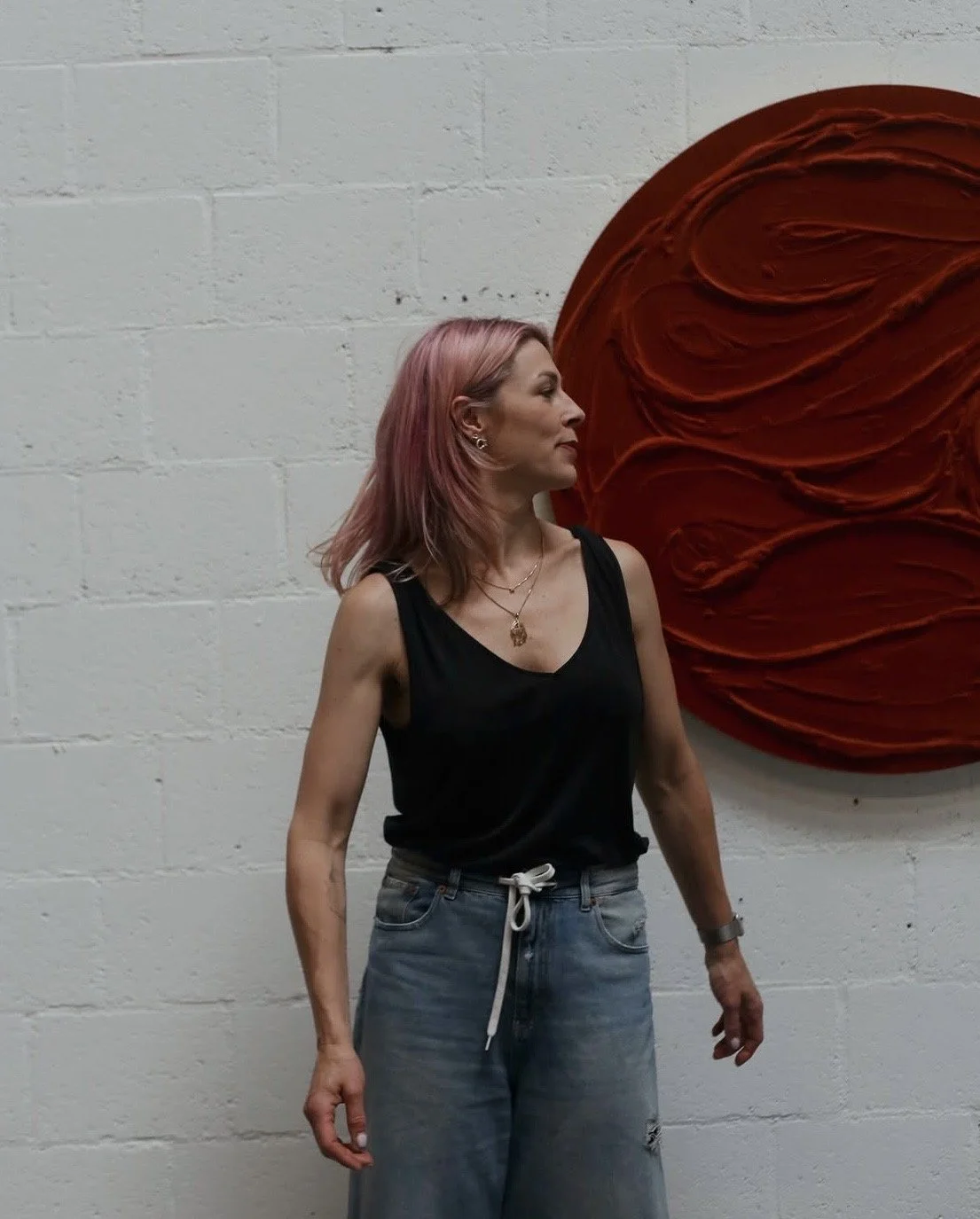 A woman with pink hair standing in profile next to a large circular red artwork on a white brick wall.