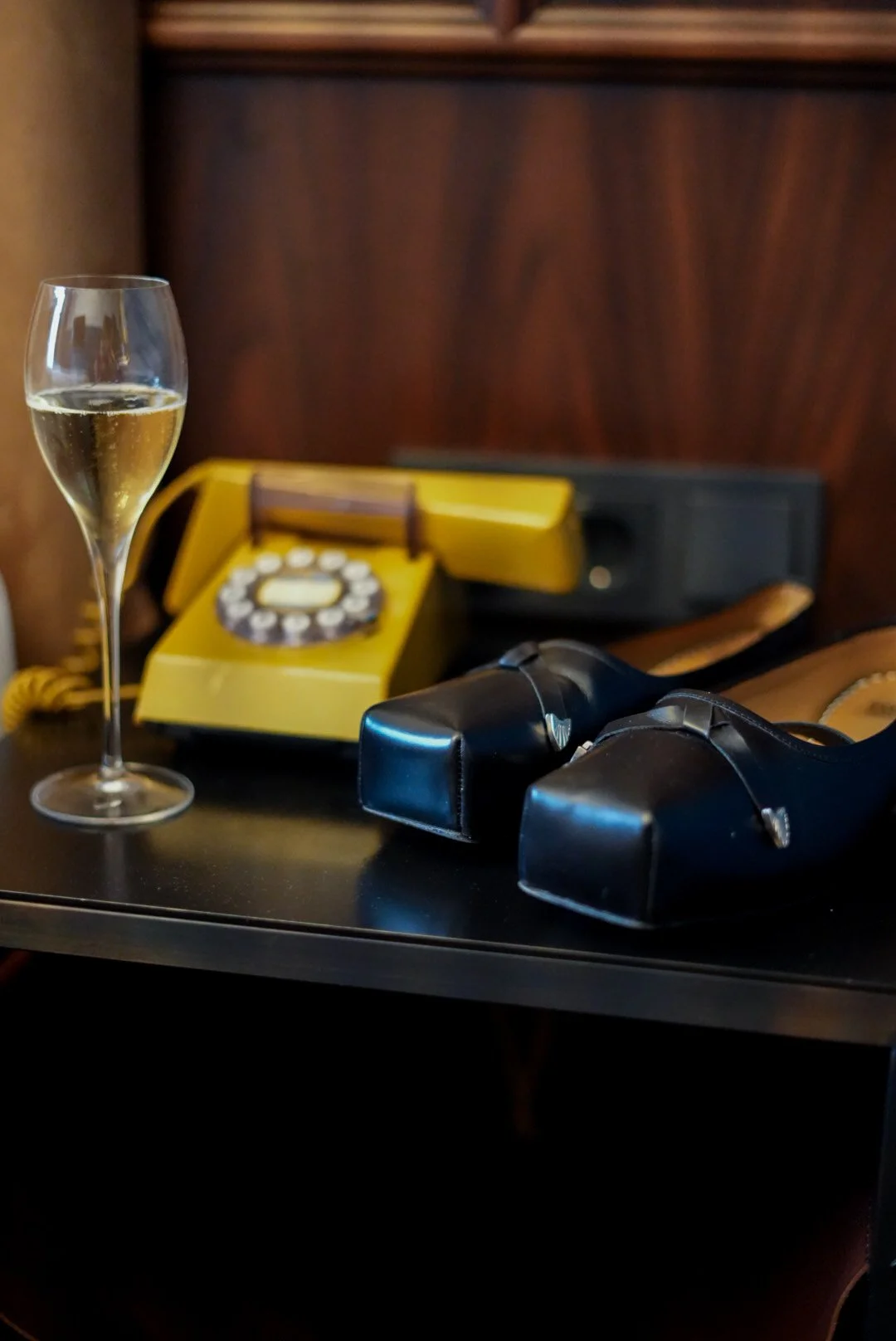 A glass of champagne, a yellow rotary phone, a pair of black high heels, and a black wallet on a dark table.