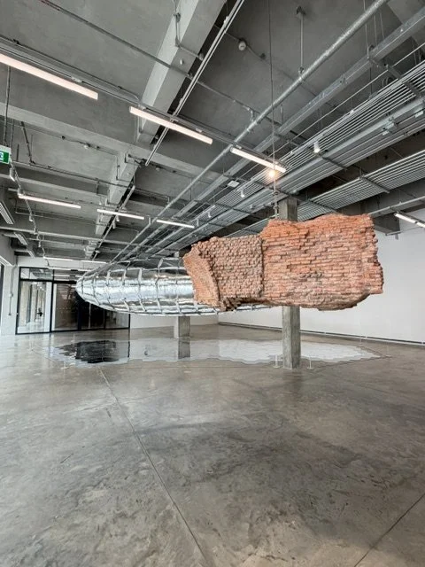An art installation featuring a large, suspended structure mimicking a section of brick wall with an industrial duct running through it, inside a modern, spacious room with a polished concrete floor and exposed ceiling pipes.