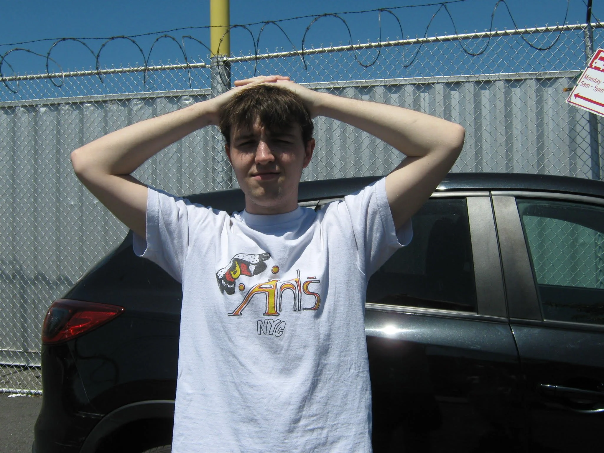Young man wearing a white T-shirt with the text "New York NYC" on front, standing outdoors with arms raised and hands resting on his head, in front of a black SUV and a chain-link fence topped with barbed wire, under a clear blue sky.