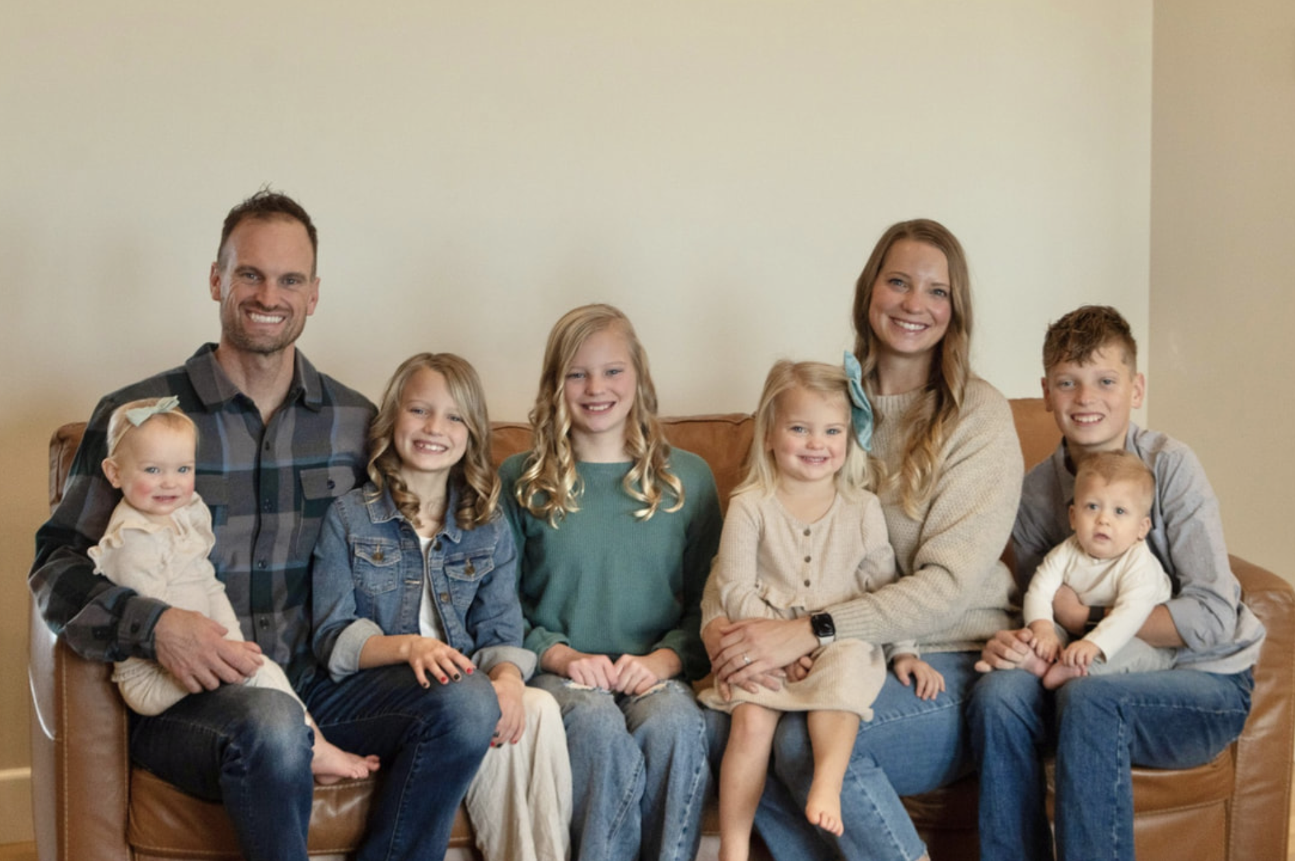 A large family sitting closely together on a brown leather couch in a living room, smiling at the camera. The group includes two adult males, two adult females, and six children of various ages, ranging from toddler to teenager. The background shows a plain off-white wall.