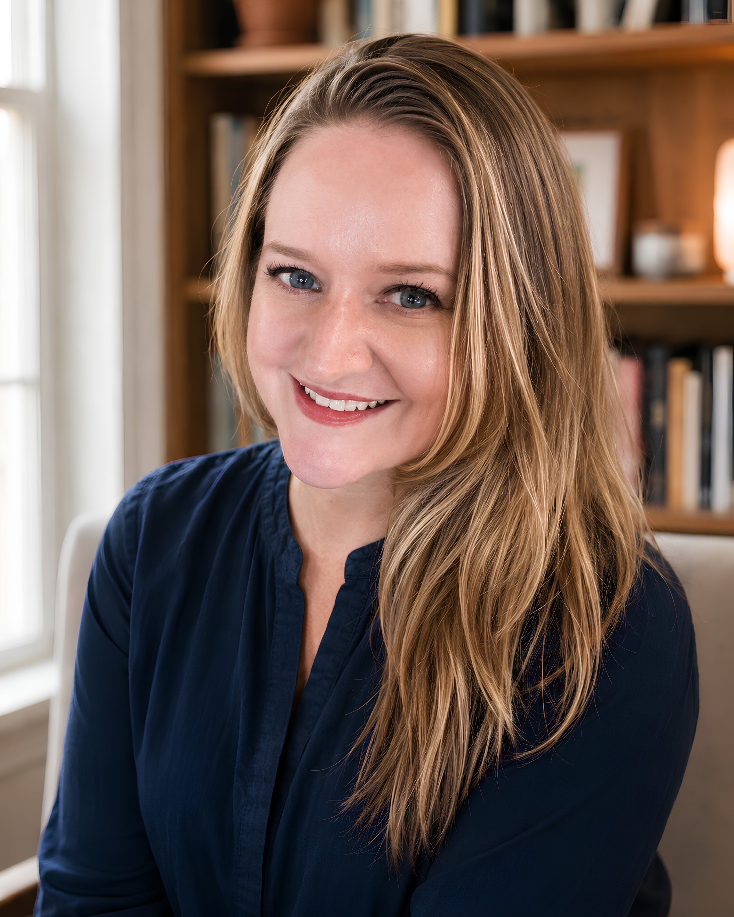 Close-up of a woman with light skin, long blonde hair, smiling, wearing a dark blue top, in a room with a wooden bookshelf in the background.