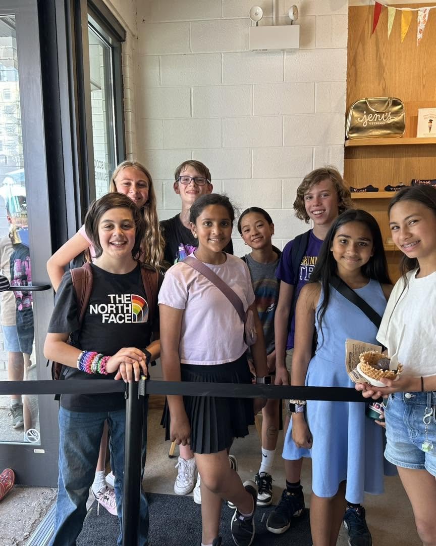 Eight diverse children standing together inside a store near an entrance, smiling for the photo. One girl on the right is holding food.