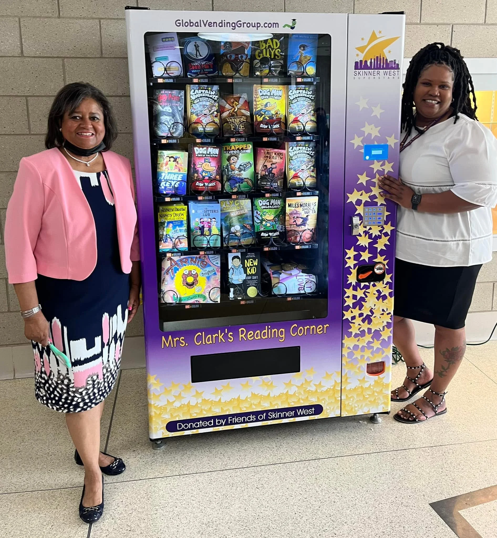 Two women standing on either side of a vending machine filled with books. The woman on the left is wearing a pink blazer over a black and white dress, while the woman on the right is wearing a white blouse with black pants and black sandals. The vending machine is labeled "Mrs. Clark's Reading Corner" and is part of a project donated by Friends of Skinner West.