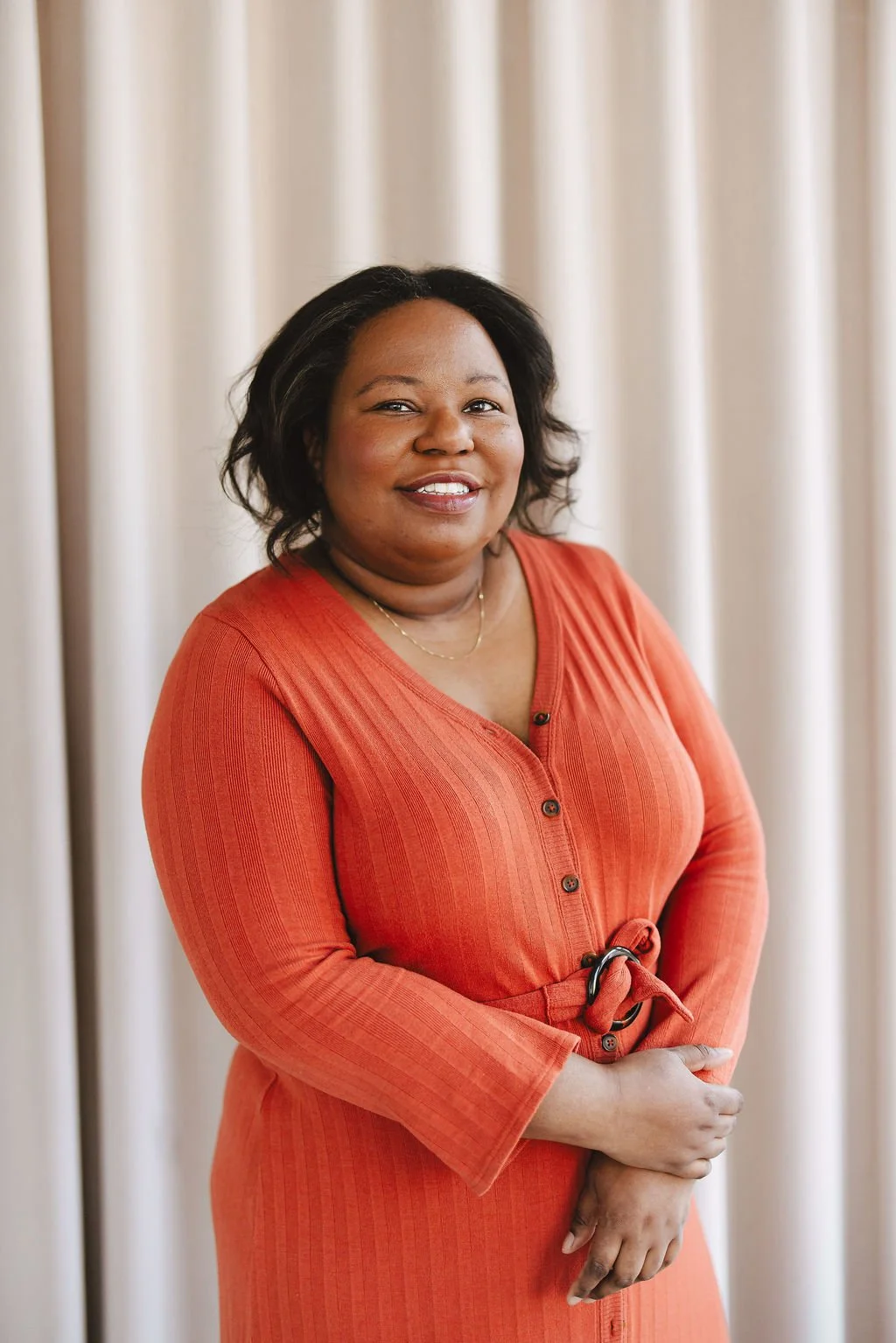 A woman with medium skin tone, shoulder-length black hair, and a warm smile, standing in front of a cream-colored curtain, wearing an orange button-down dress with a belt.