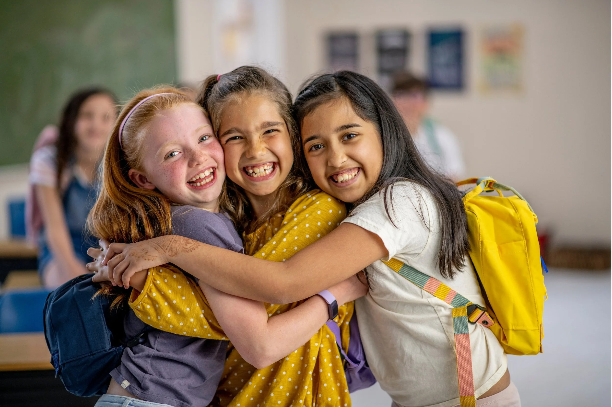 Three diverse school girls are hugging and smiling in a classroom, showing friendship and happiness.