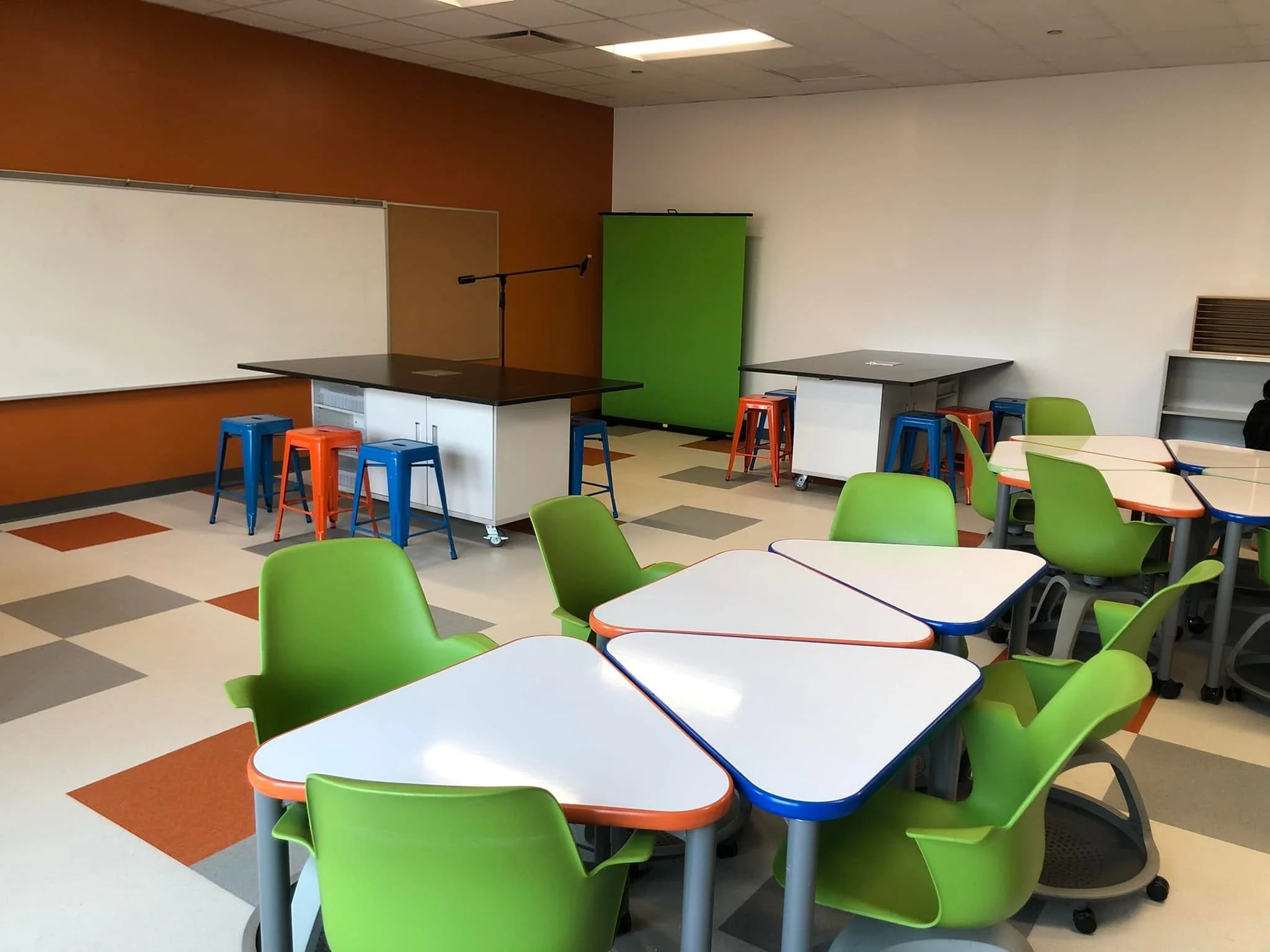 An empty classroom with bright green chairs and white tables with orange and blue edges. The room has a whiteboard, a green screen, and a speaker's podium, with a ceiling light illuminating the space.