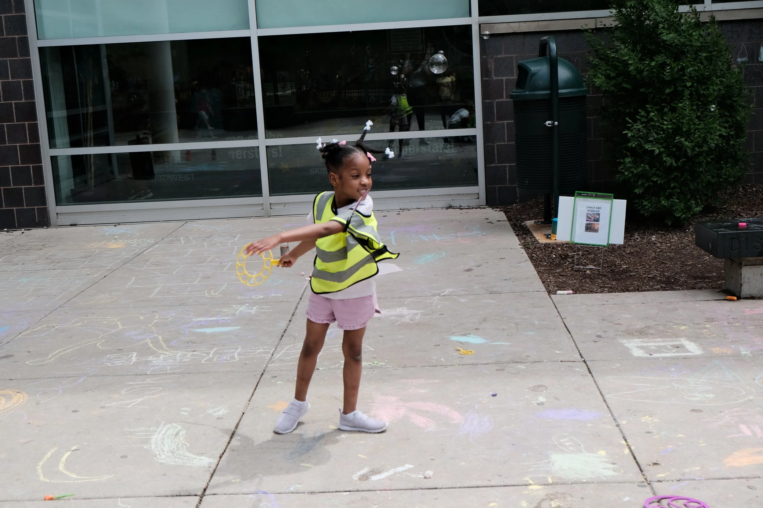A young girl with braided hair in buns, decorated with pink and white hair accessories, is joyfully spinning around holding a yellow bubble wand, outside in front of a glass building with chalk drawings on the sidewalk and a small chalkboard sign nearby.