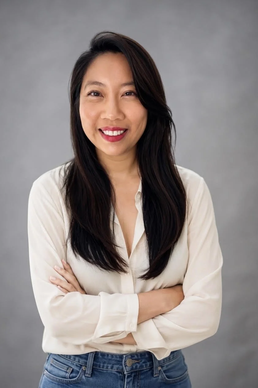 A woman with long black hair smiling and crossing her arms, wearing a white shirt and jeans, in front of a gray background.