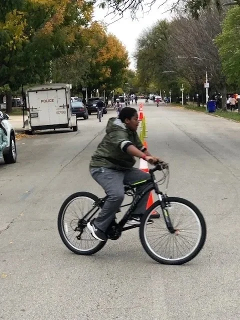 A boy riding a black bicycle on a residential street with autumn-colored trees and traffic cones.