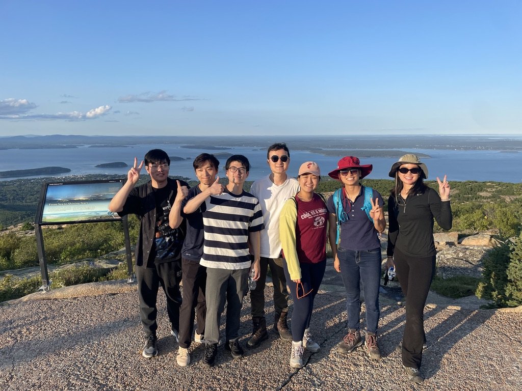 Group of eight people posing outdoors with a scenic view of a lake and hills in the background, some making peace signs.