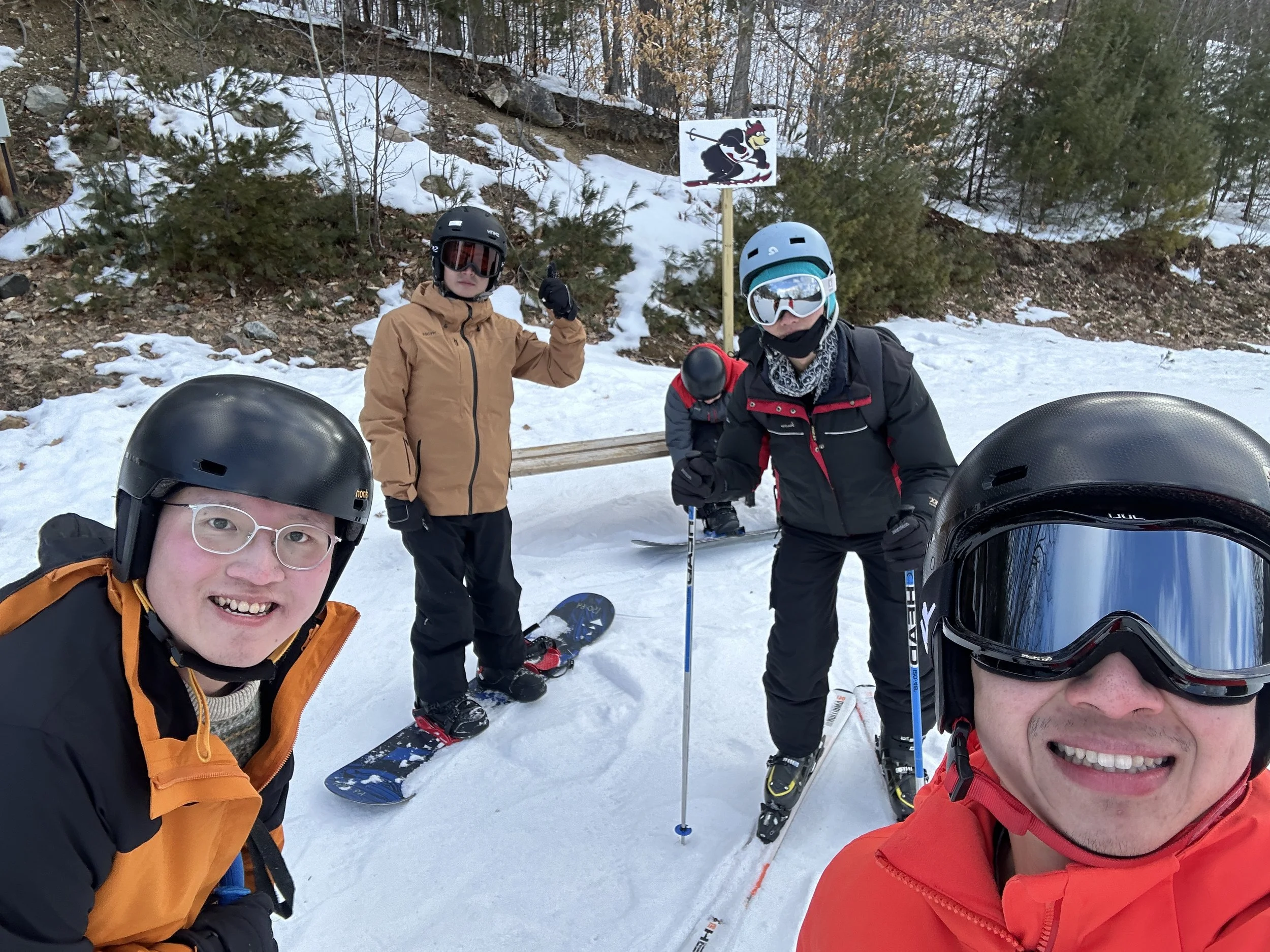 Group of five skiers in winter gear and helmets on snowy mountain with trees and a sign showing a cartoon dog with skis.