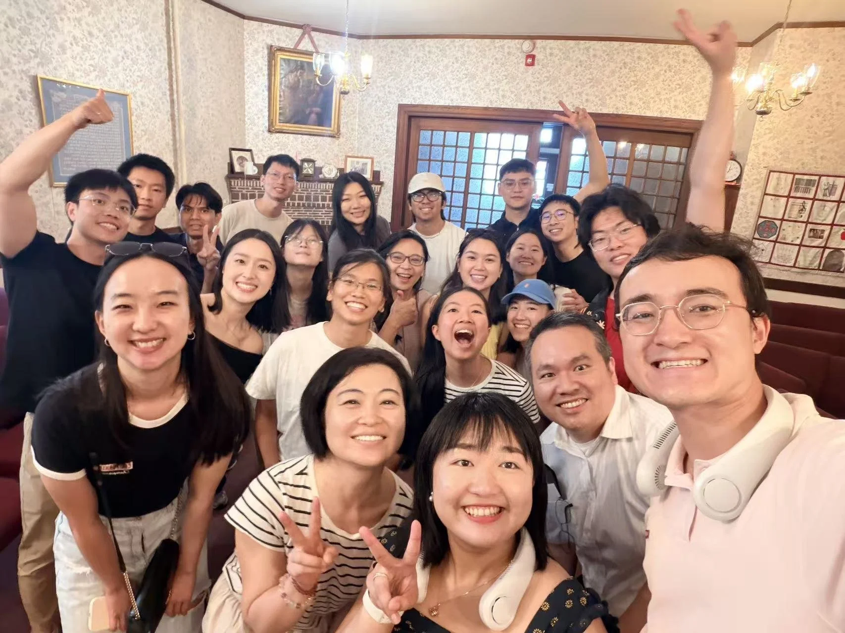 A large group of people smiling and making peace signs in a cozy room with floral wallpaper, artwork, and warm lighting.