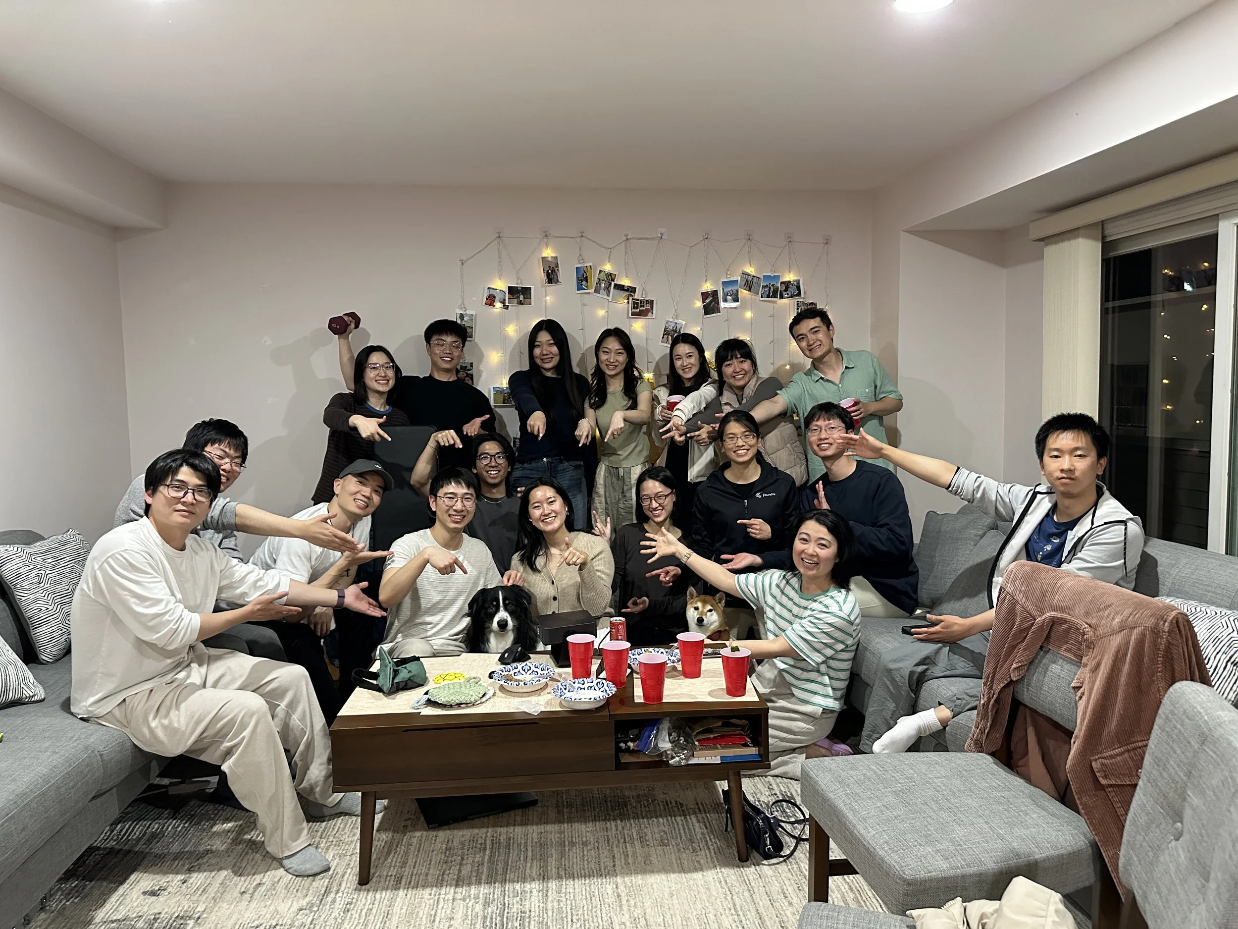 A group of people gathered in a living room celebrating, with some sitting on sofas and others standing. The room is decorated with string lights and photos hung on the wall. There are snacks and drinks on the table, and two dogs are present in the s