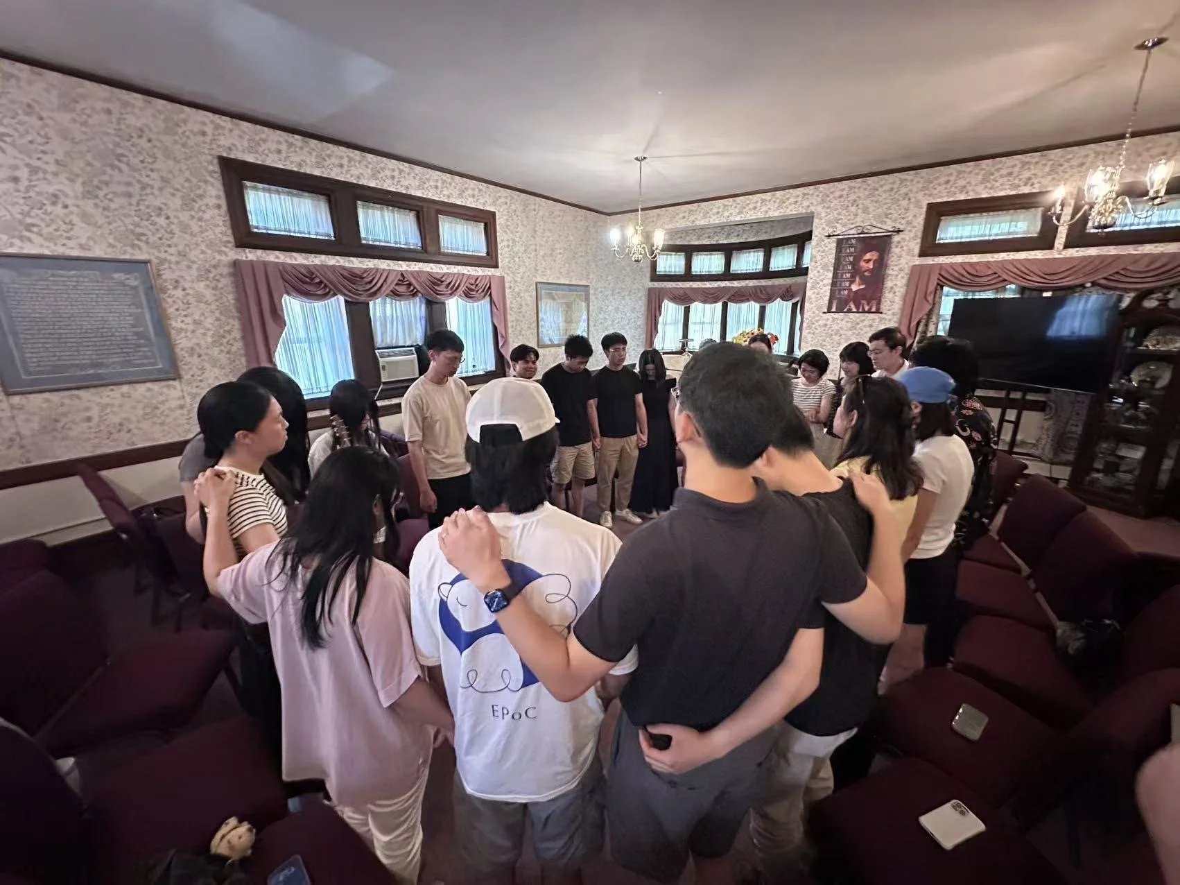 Group of people standing in a circle with their heads bowed and arms linked in a prayer or moment of reflection in a well-lit living room with floral wallpaper, curtains, and a chandelier.