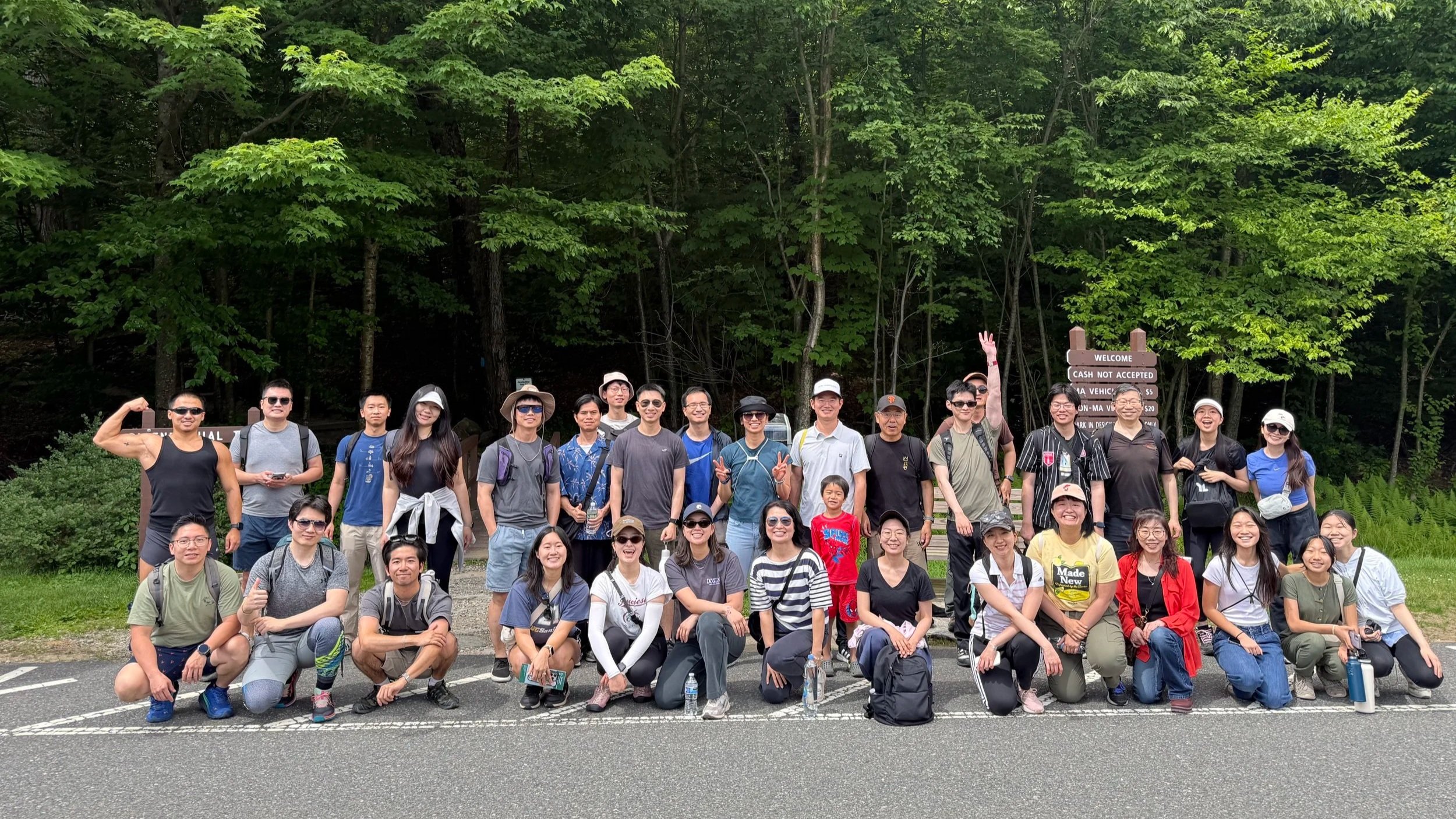 Group of diverse people outdoors, smiling and posing for a photo with trees and a sign in the background.