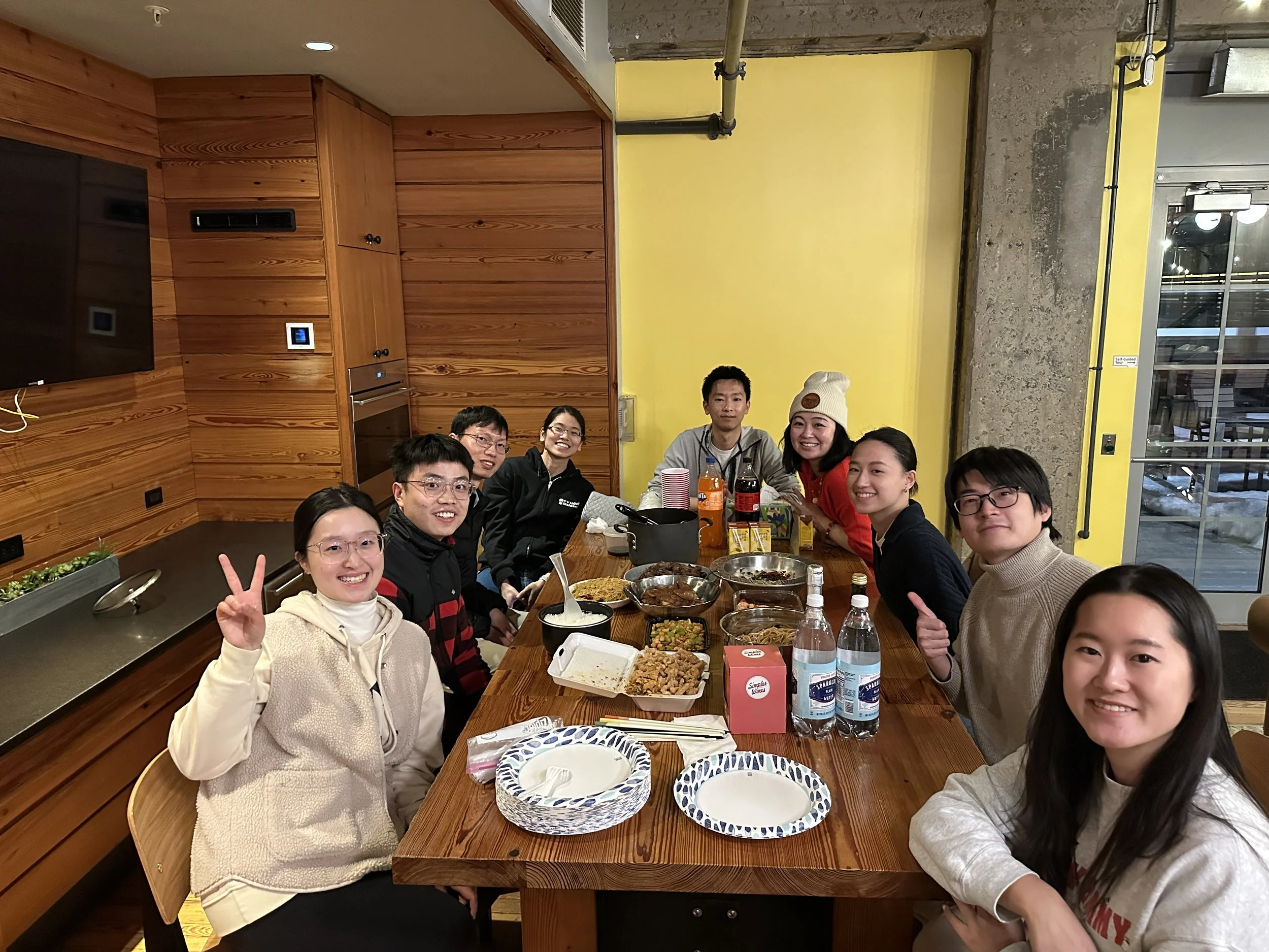 A group of people sitting around a dining table enjoying a meal together at a restaurant. There are various dishes and drinks on the table, and some people are smiling and making peace signs.