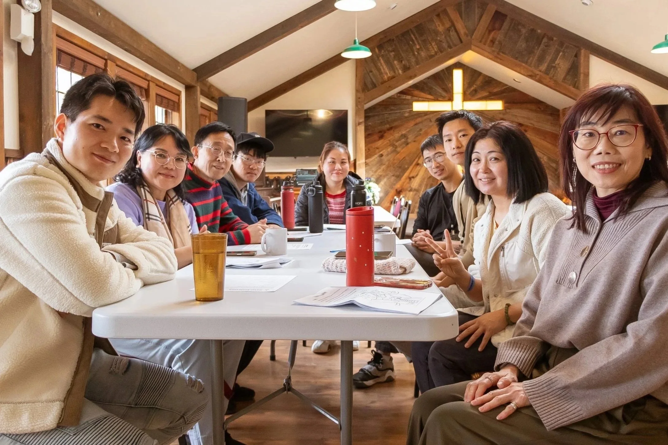 A group of nine people sitting at a long table inside a room with wooden walls and ceiling, with books, papers, water bottles, and mugs on the table, smiling and looking at the camera.