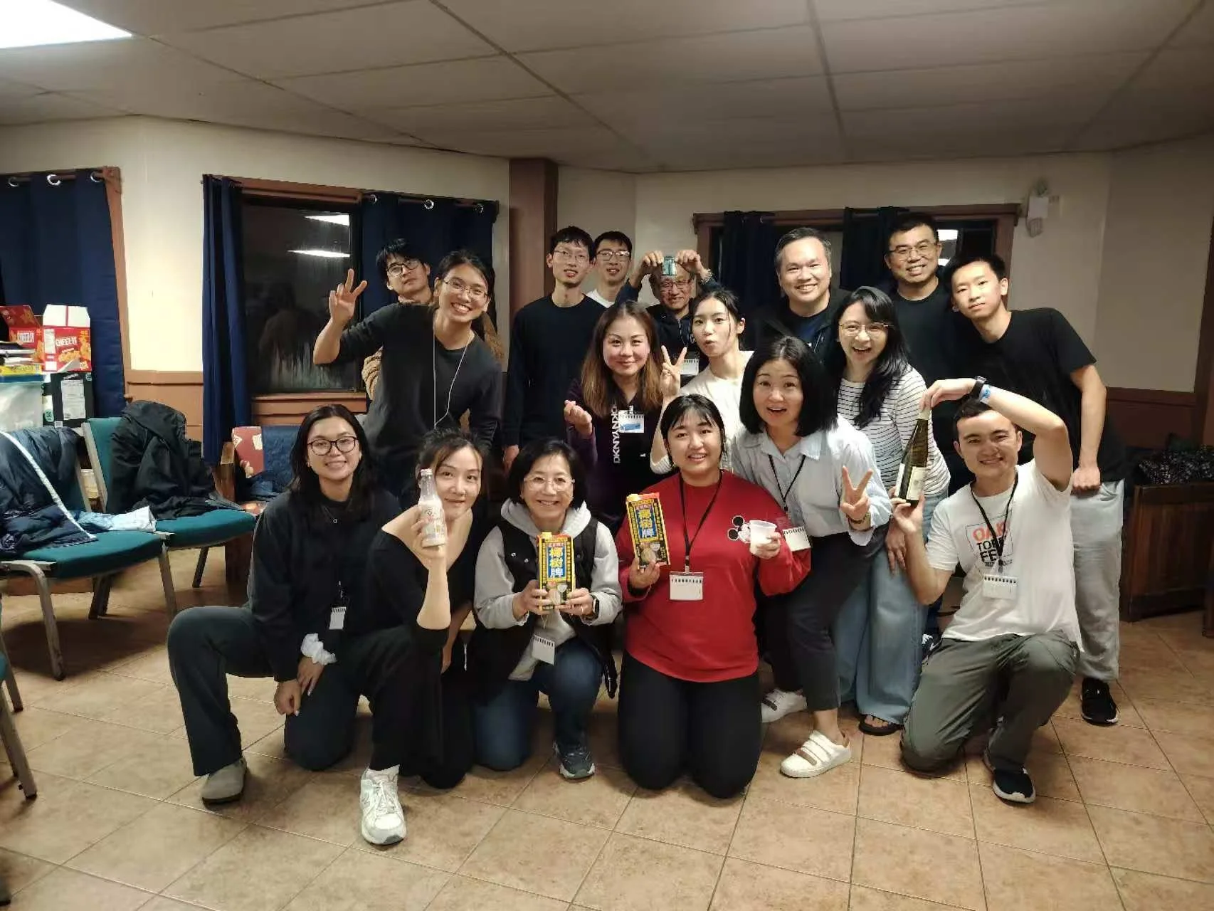Group of people smiling and posing for a photo in a room with beige tiled floors, dark blue curtains, and a wooden wall. Some are holding snacks and drinks, and the group includes men and women of East Asian descent.