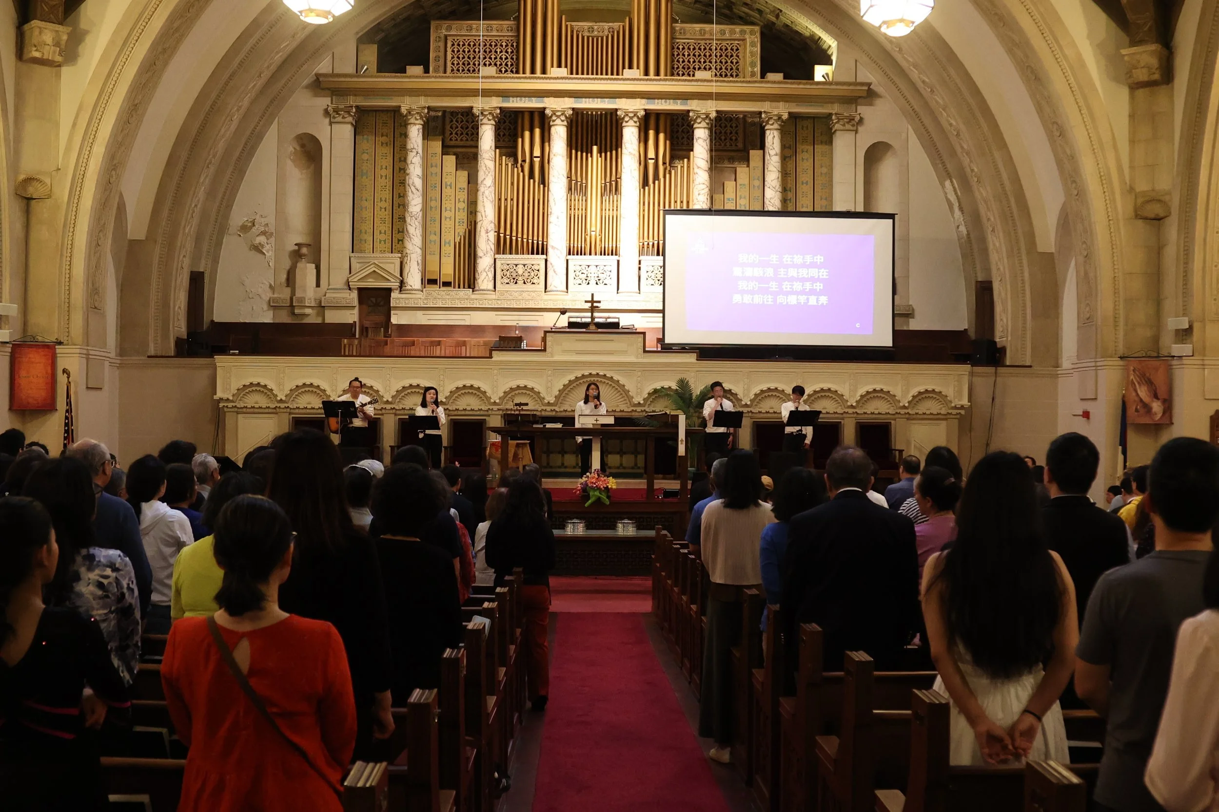 A church interior with a congregation, a stage with a cross, and choir members singing with a large screen displaying text in Chinese.