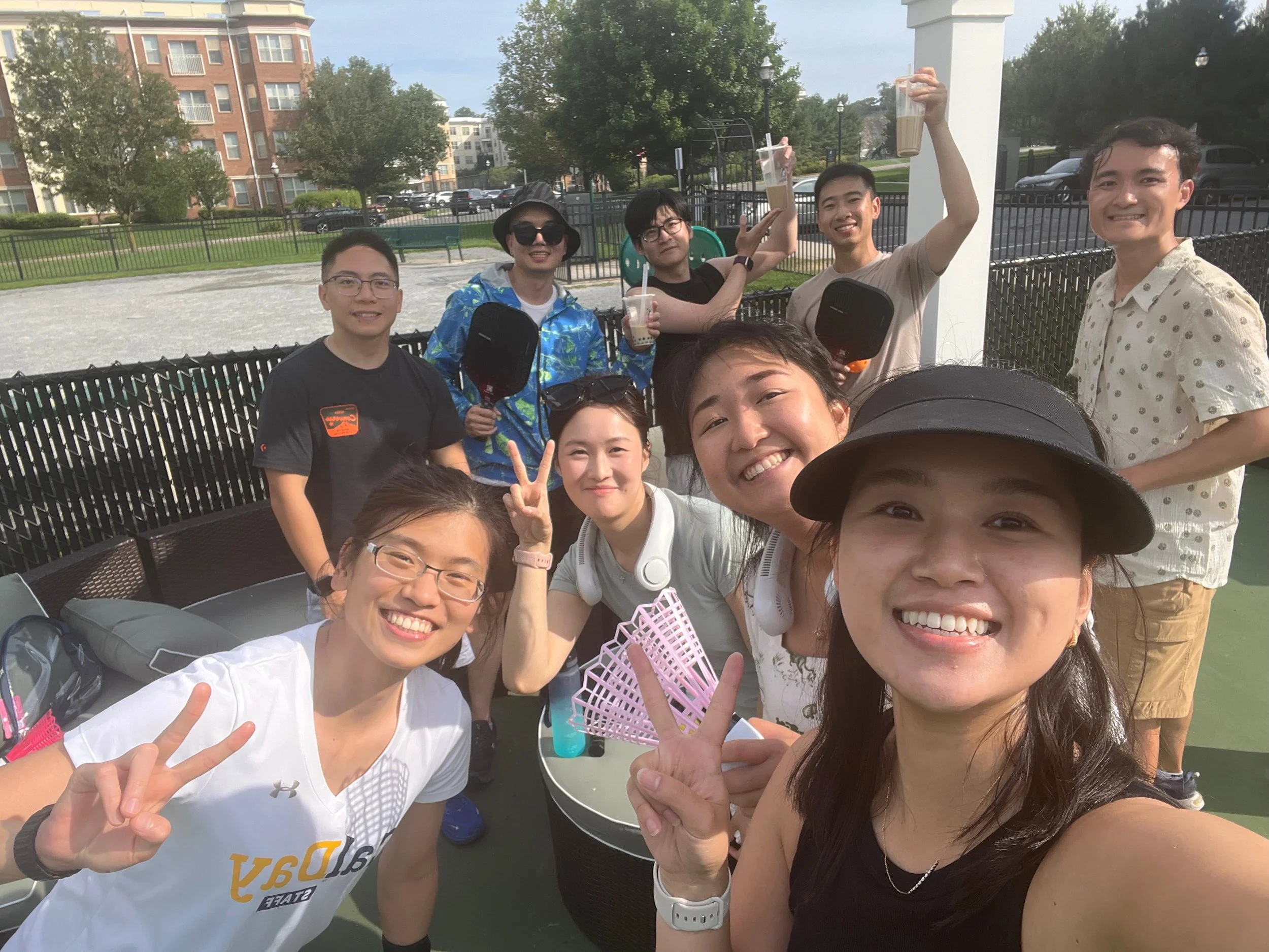 A group of smiling young adults posing outdoors on a sunny day, some making peace signs and holding drinks, with apartment buildings and trees in the background.