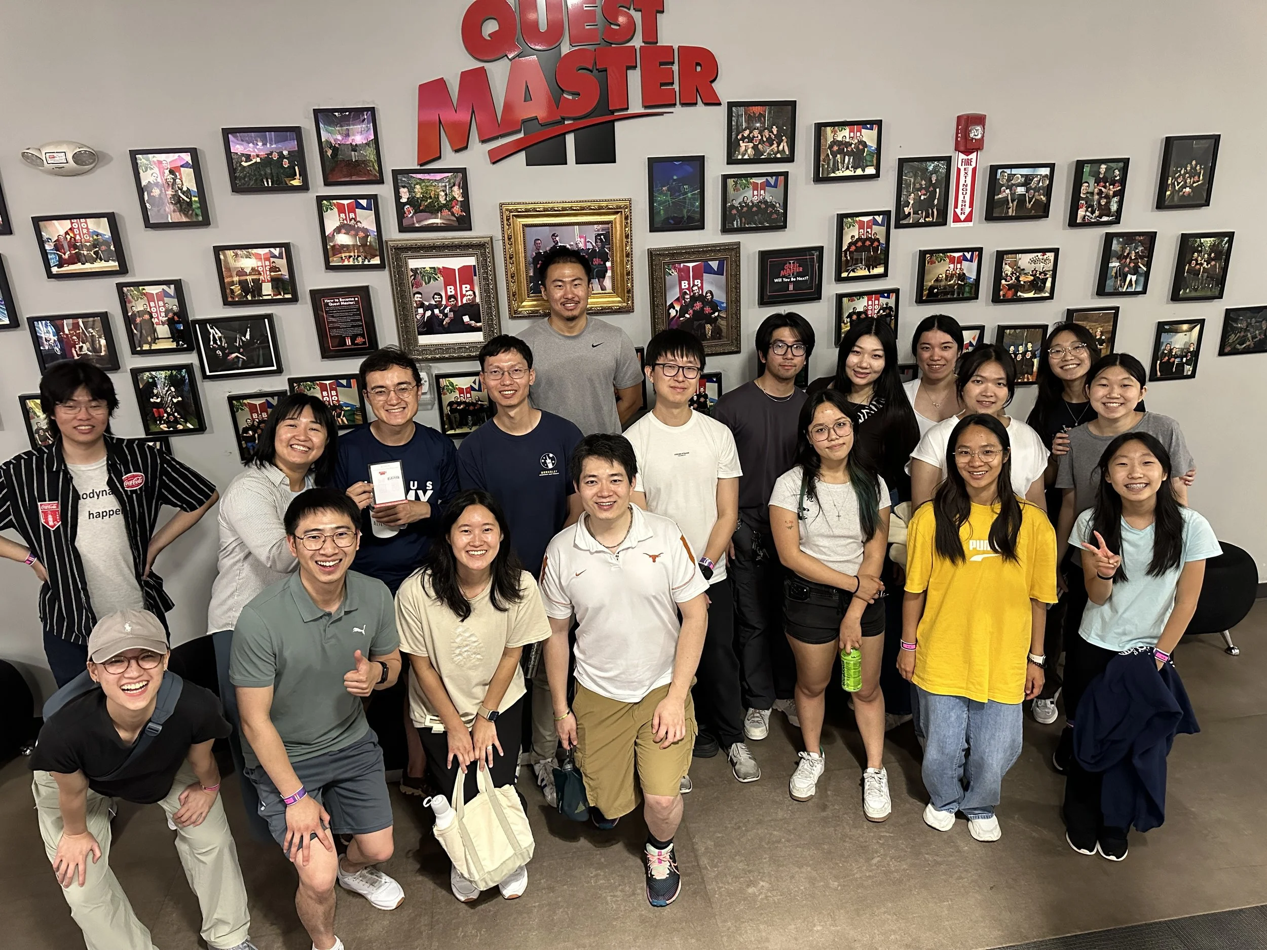 A group of diverse young adults posing for a photo in front of a wall decorated with numerous framed photographs and a large red sign that reads 'Quest Master.' The group appears cheerful, with many smiling at the camera.
