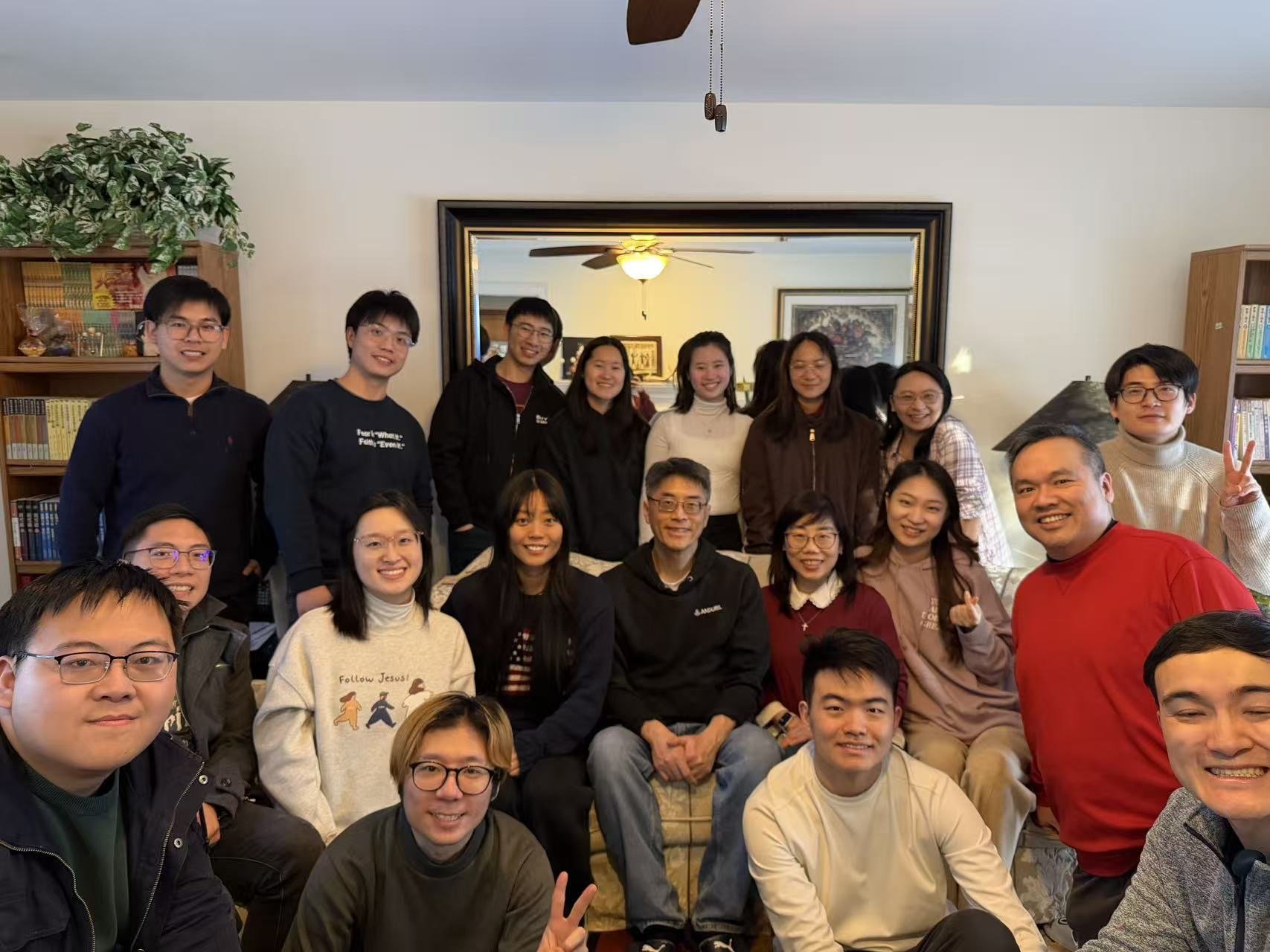 Group of people smiling and posing for a photo in a living room, with a large mirror, bookshelves, and a ceiling fan in the background.