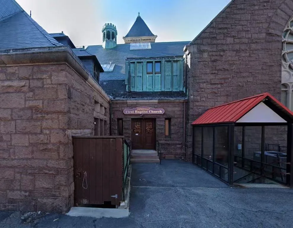 Entrance of the First Baptist Church with a wooden door, stone brick walls, and a small staircase, with a protective glass shelter on the right side.