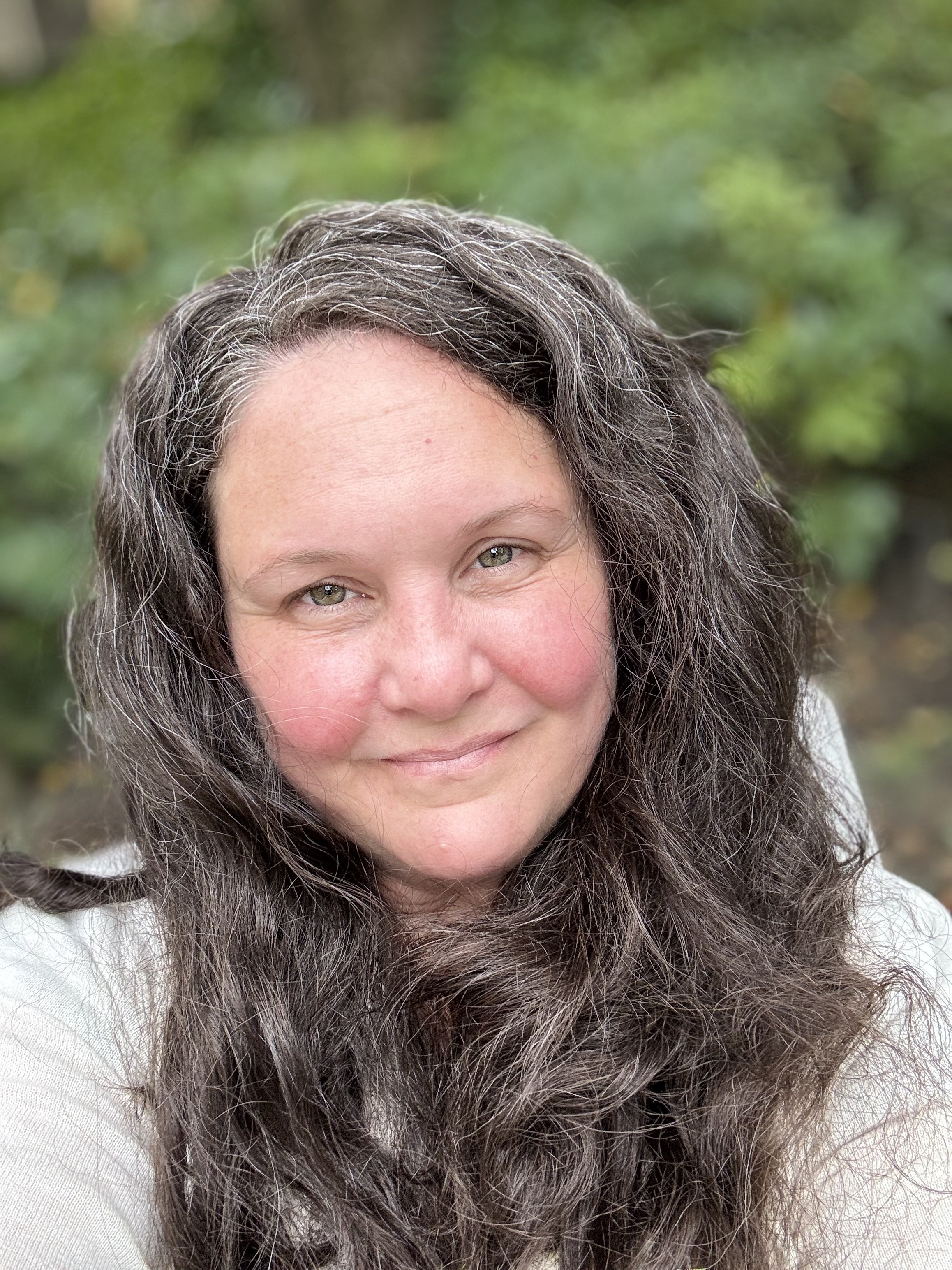 Close-up of a woman with gray curly hair, smiling, outdoors with green leafy background.