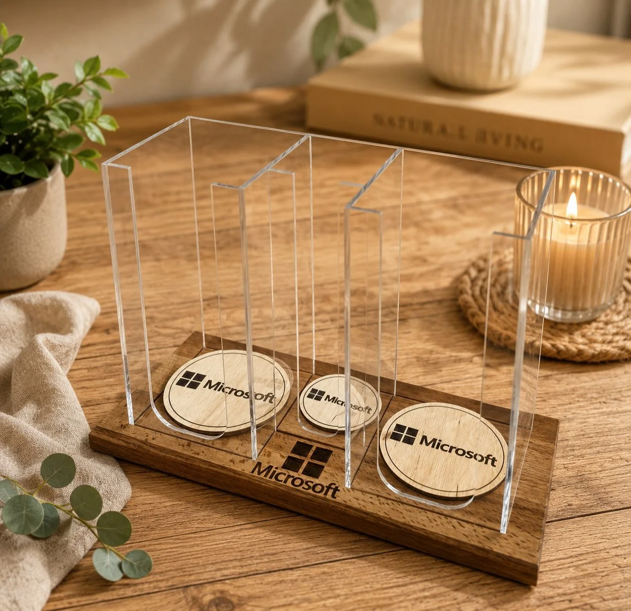 A transparent acrylic award with three sections on a wooden base, featuring the Microsoft logo on circular wooden plaques. The award is placed on a wooden table next to a potted plant, a white cloth, and a lit candle in a glass holder on a woven coaster. In the background, a book titled 'Natural Living' is visible.