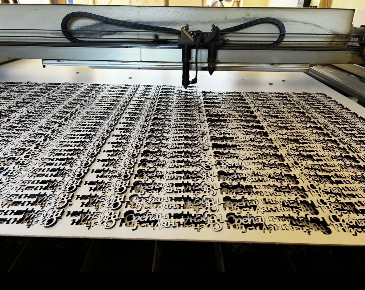 A laser cutting machine producing a sheet with repeated cutout words that say "Hey" and "Haha" in black on a white background.